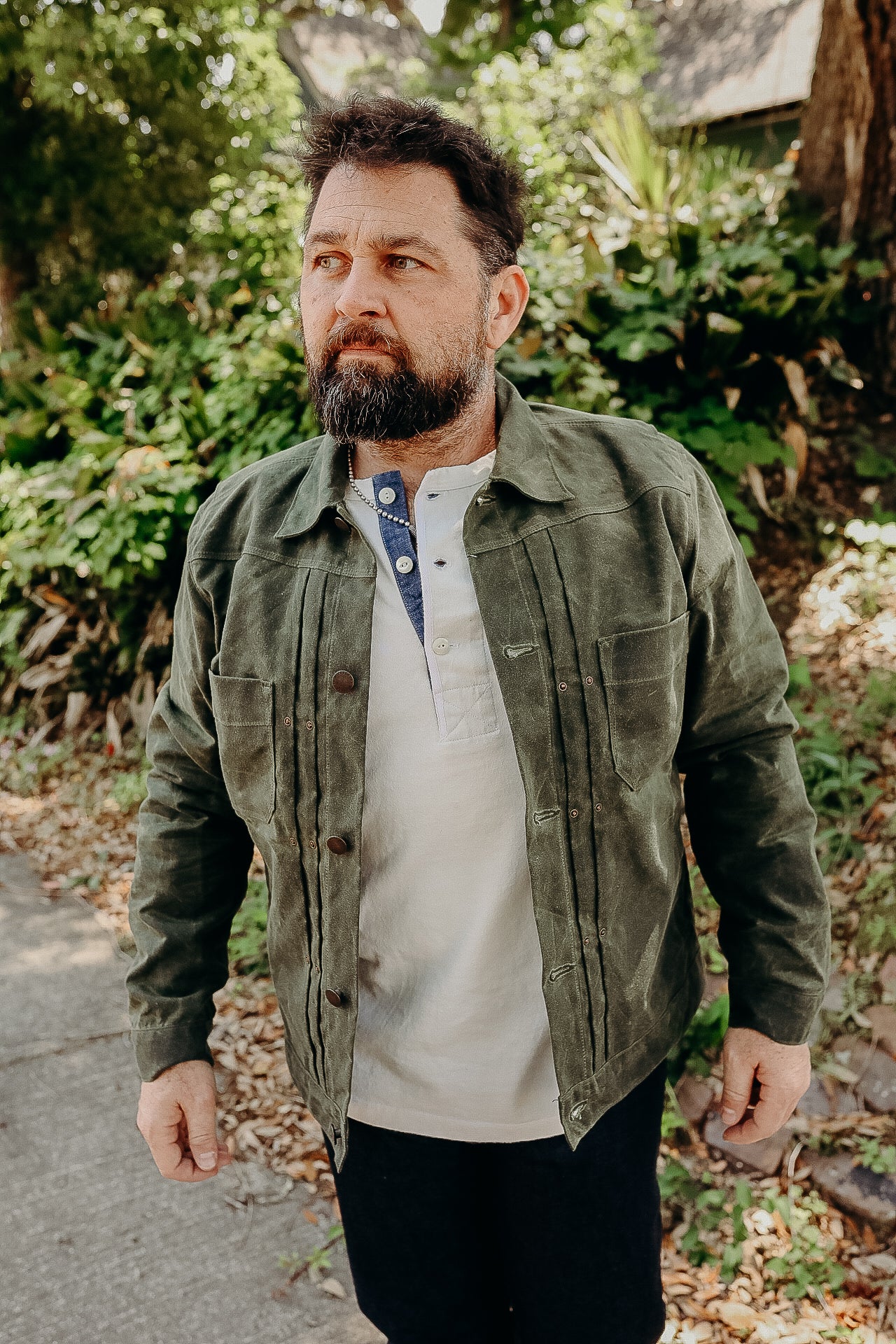 Man wearing olive green waxed cotton work jacket over white henley shirt outdoors with leafy background
