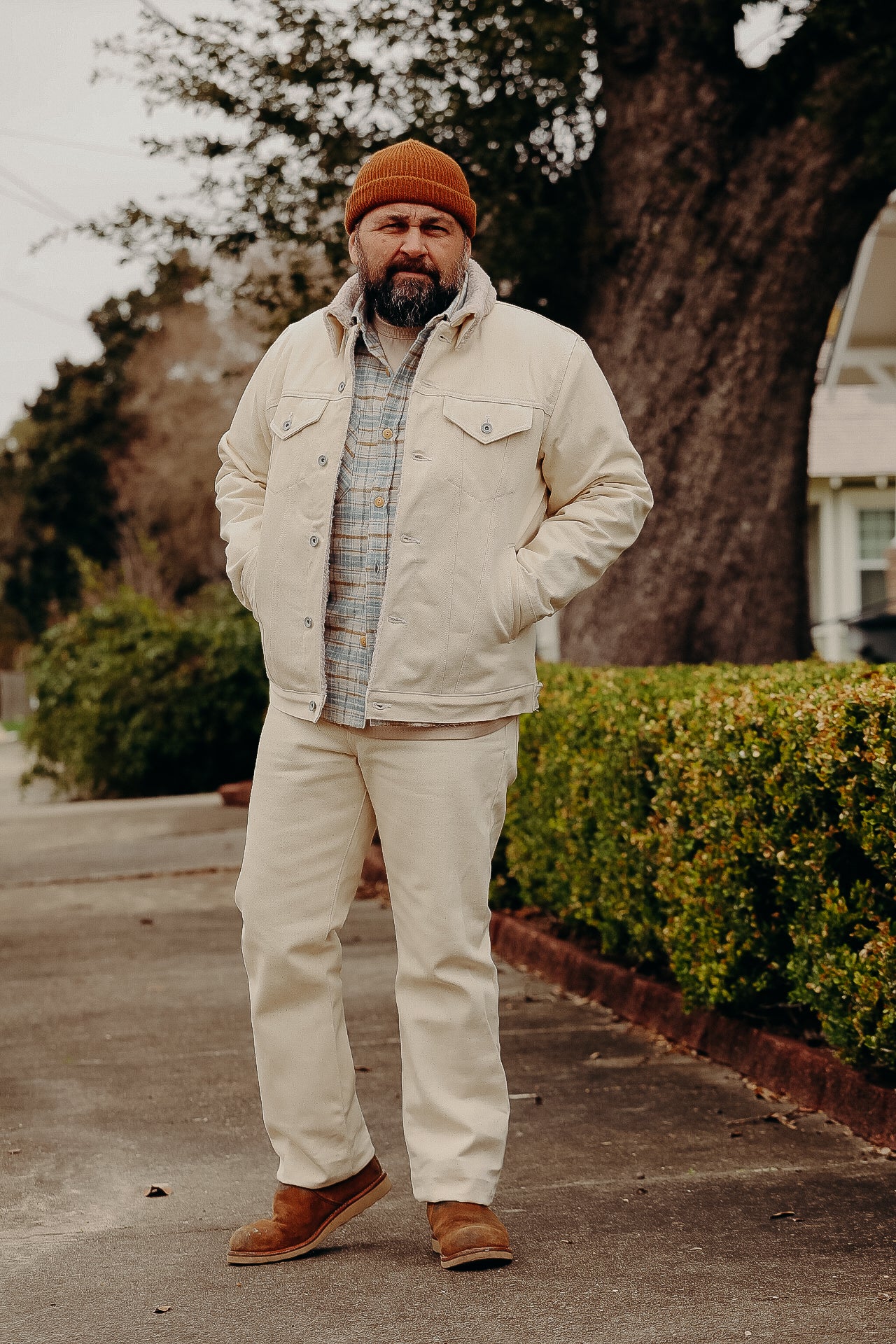 Man wearing cream denim jacket, beige pants, plaid shirt, brown boots, and orange knit beanie, standing on sidewalk