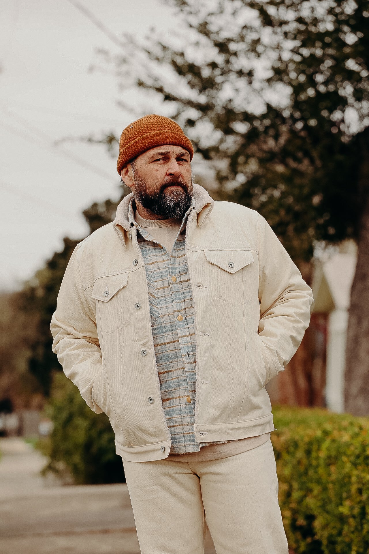Bearded man wearing a tan beanie, cream jacket, and plaid shirt, posing outdoors on a street