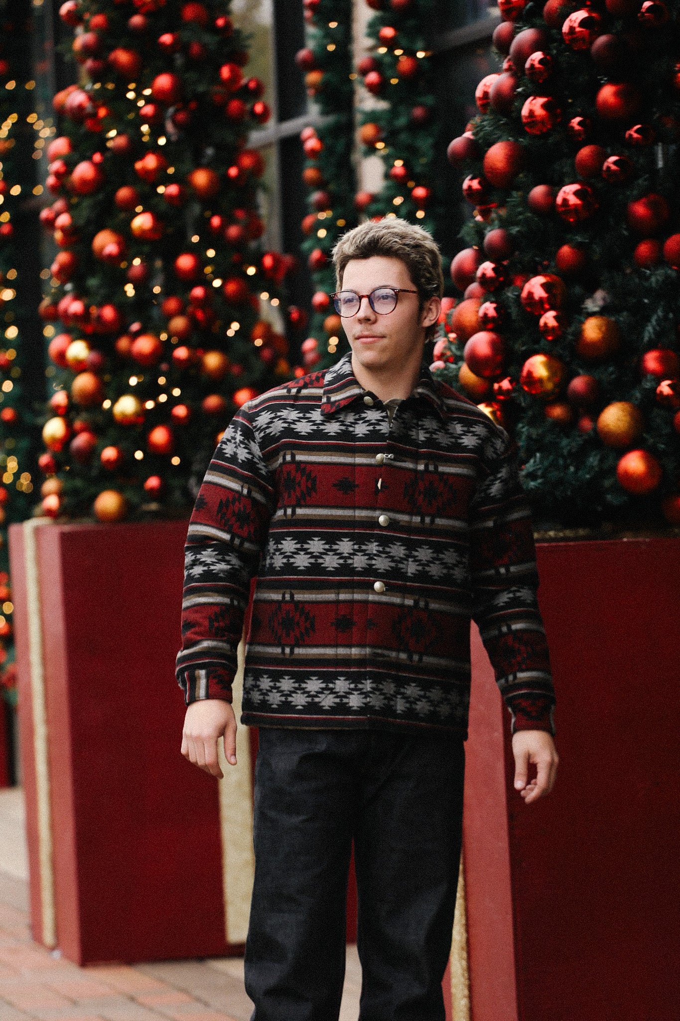 Man wearing patterned jacket and glasses standing outside near Christmas trees decorated with red ornaments and lights