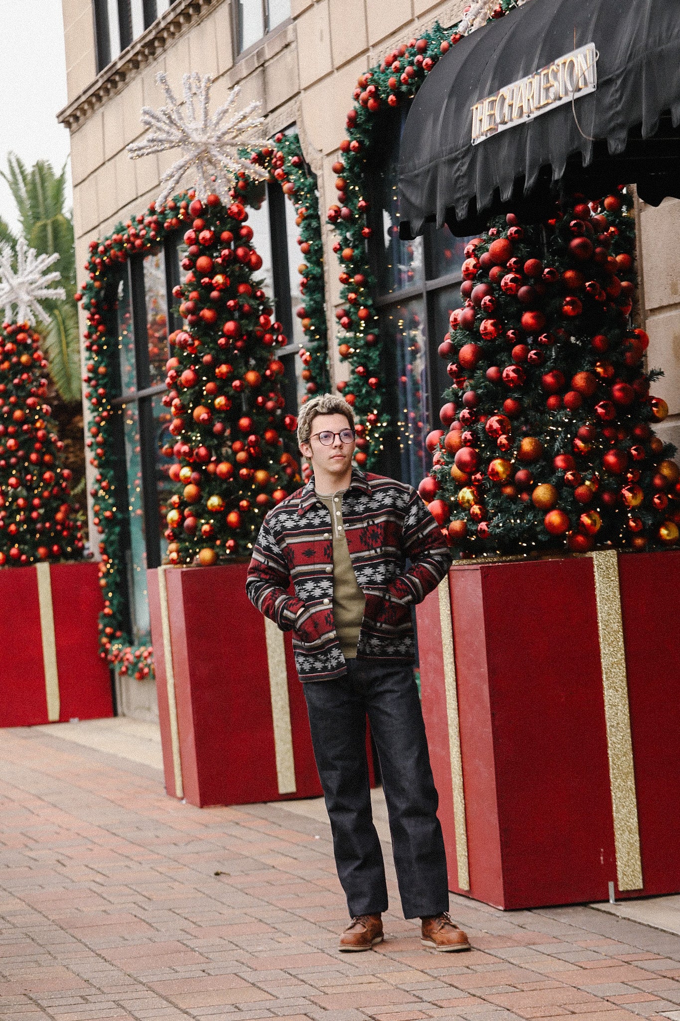 Man wearing patterned jacket and glasses standing by holiday decorated storefront with red and gold Christmas trees