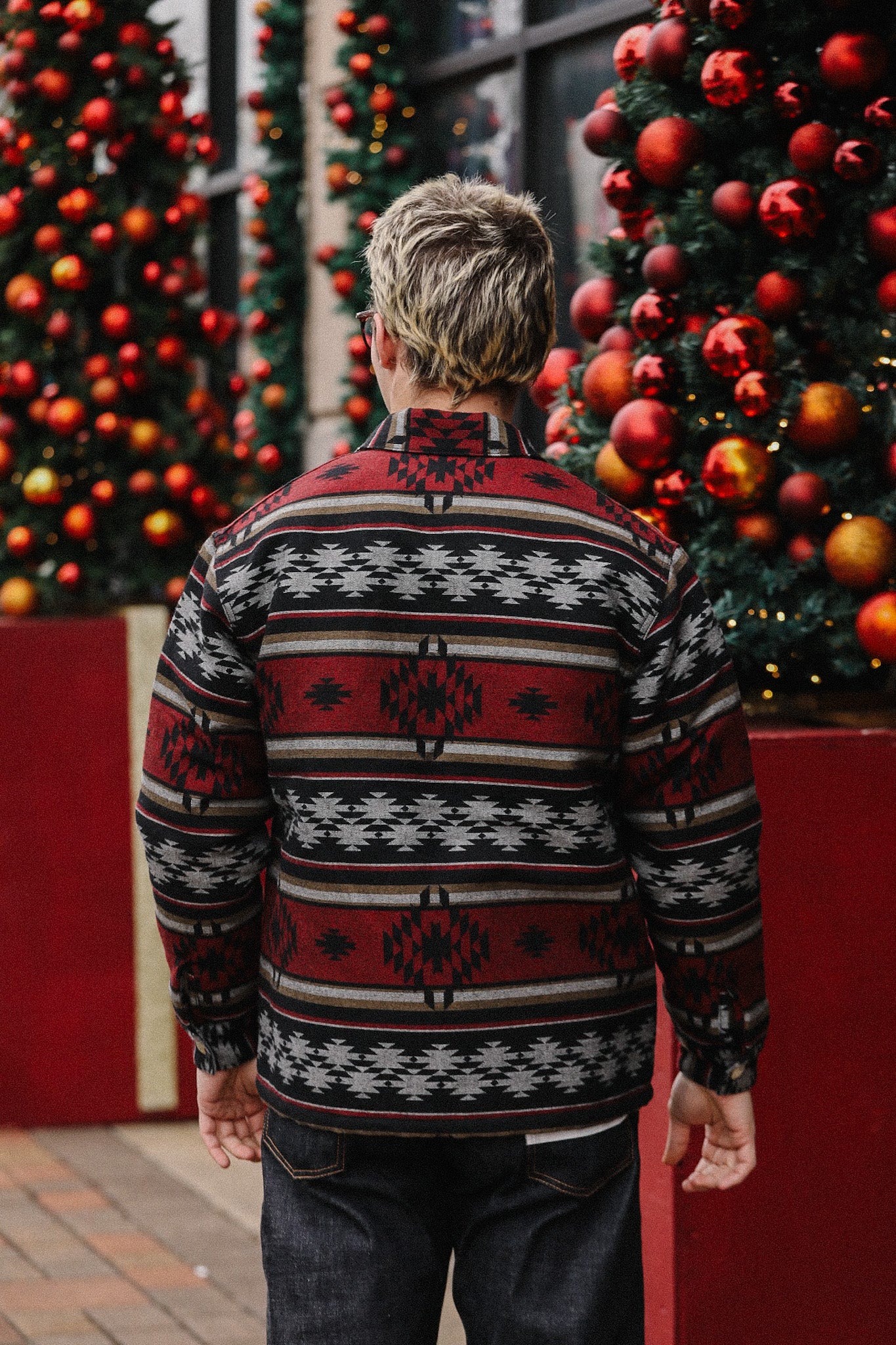 Man wearing red and black patterned jacket standing outdoors near decorated Christmas trees with red ornaments