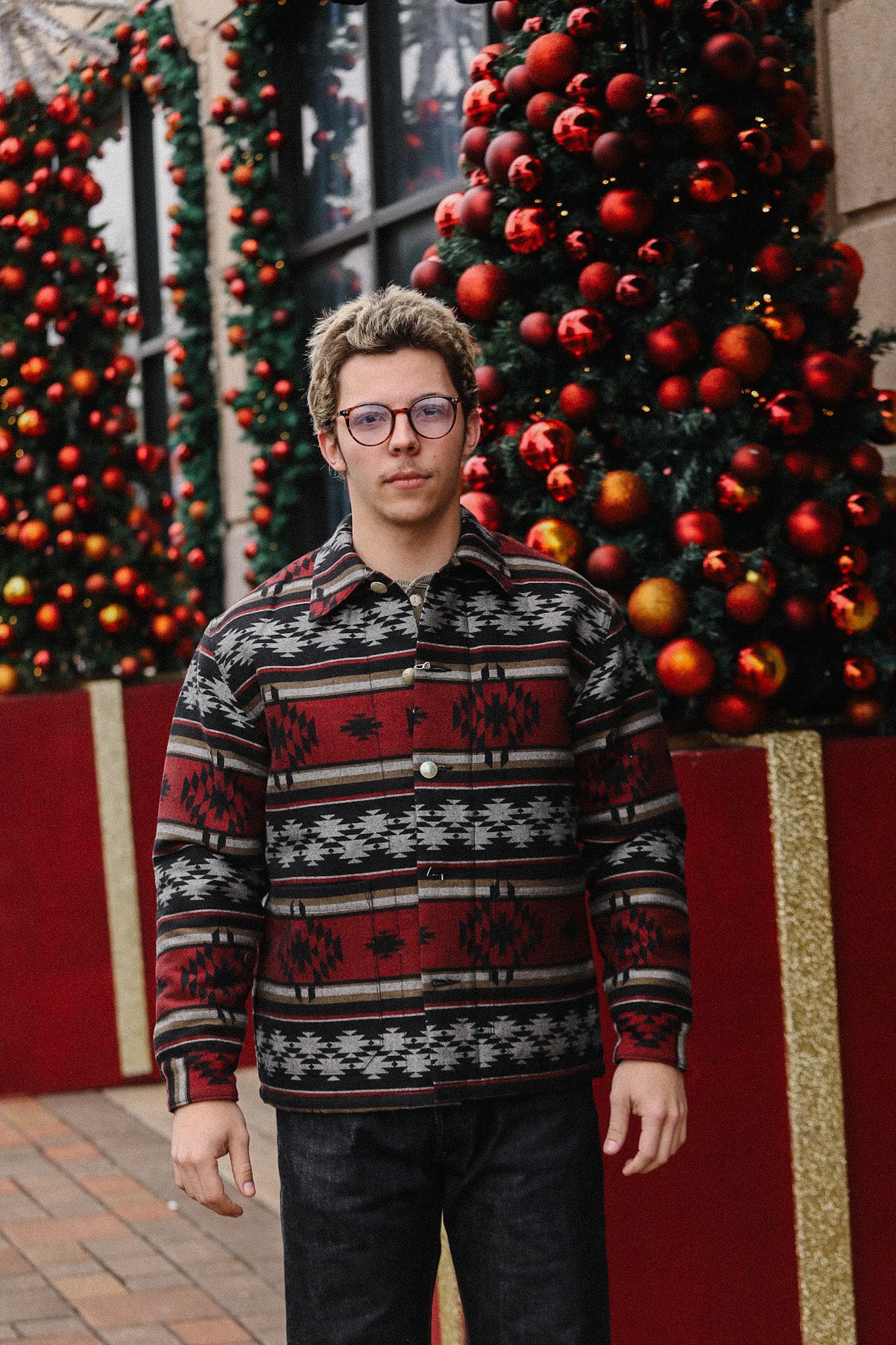 Young man wearing patterned red and black jacket standing in front of large Christmas trees decorated with red ornaments