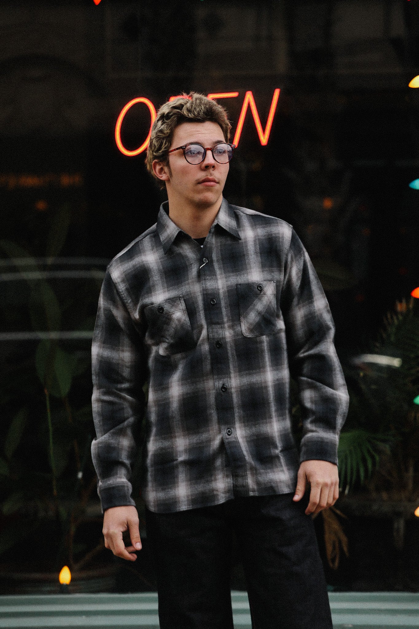 Young man wearing black and gray plaid shirt with glasses standing outdoors near neon open sign