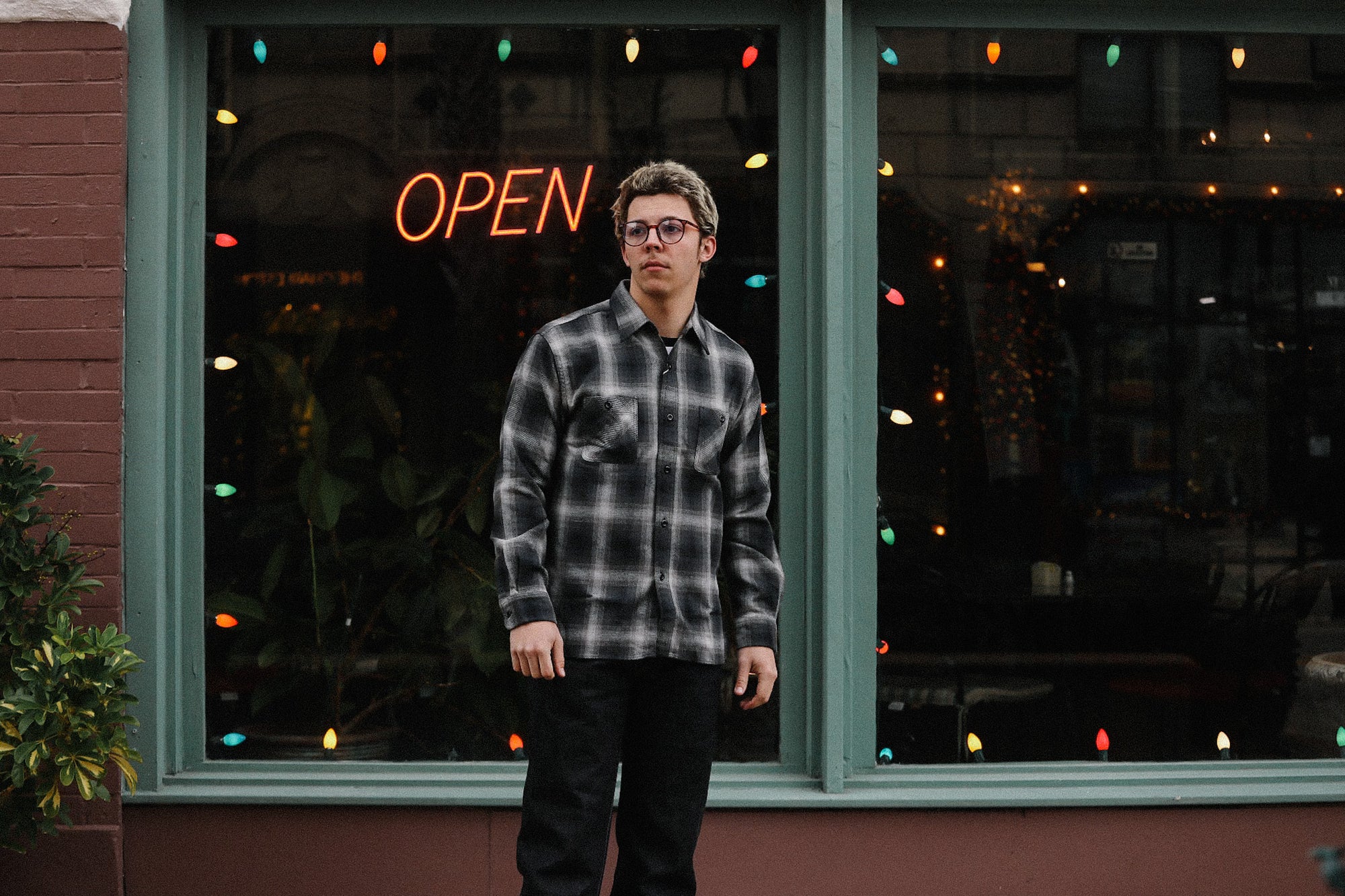 Young man wearing black and white plaid shirt standing outside a storefront with neon open sign and colorful lights