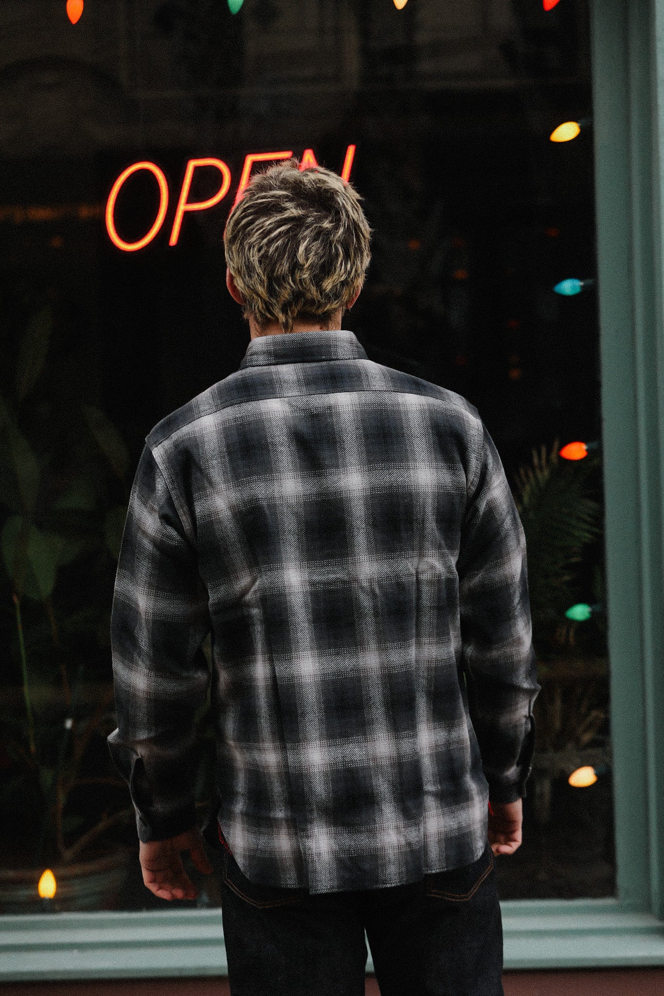 Person wearing black and gray plaid shirt standing in front of a store window with red neon open sign