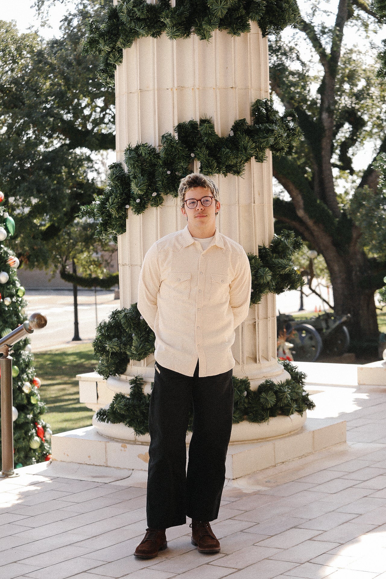 Man wearing a cream shirt and black pants standing in front of a decorated column with green garlands outdoors