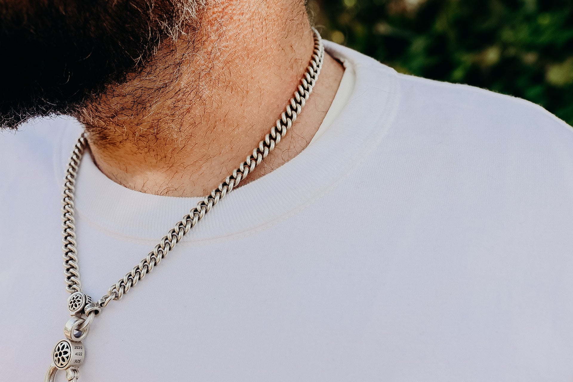 Close-up of a man wearing a thick silver chain necklace with decorative pendants over a white shirt
