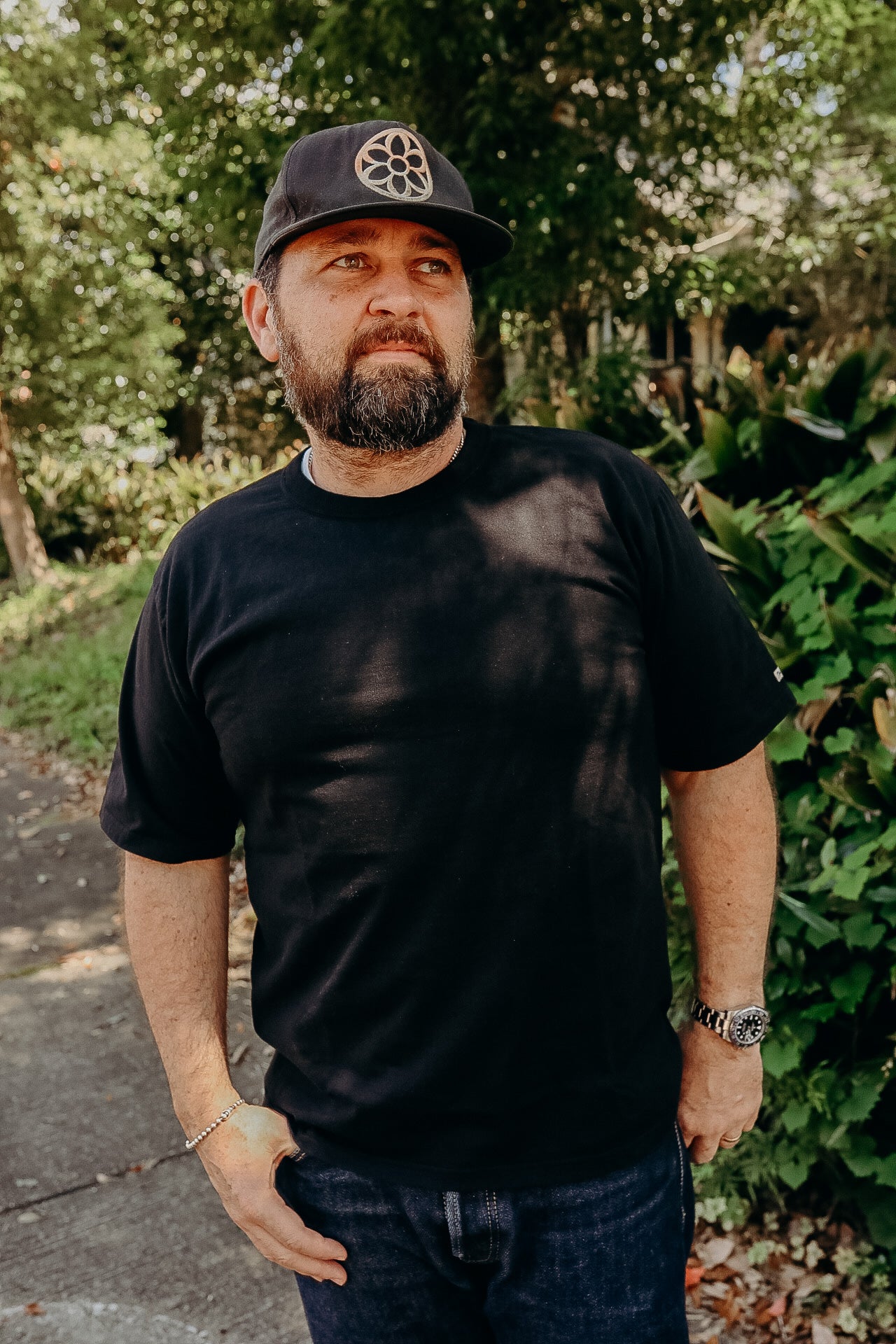 Man wearing black cap with floral logo, black t-shirt, and wristwatch standing outdoors near green foliage