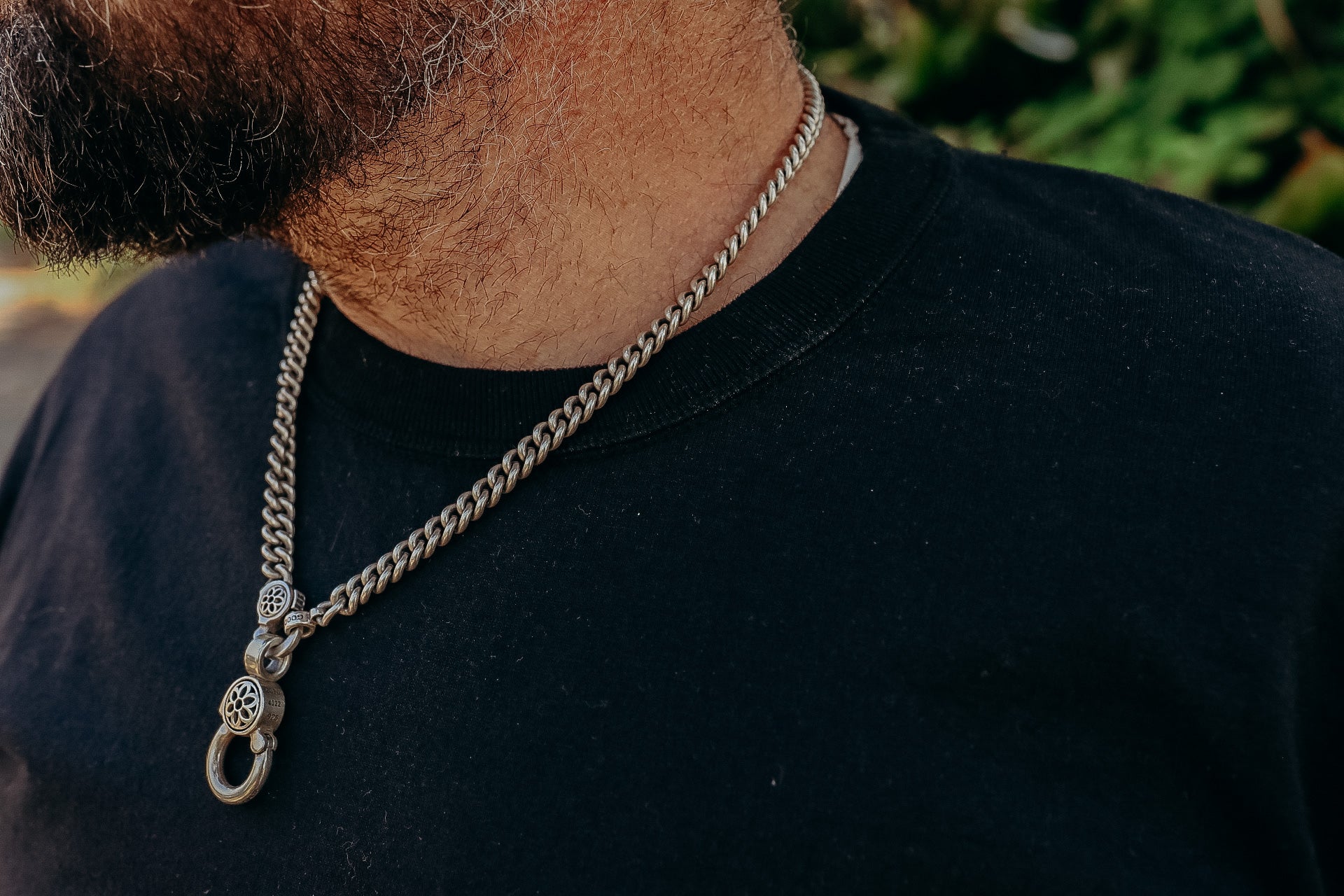 Close-up of a man wearing a silver chain necklace with detailed clasp over a black shirt outdoors