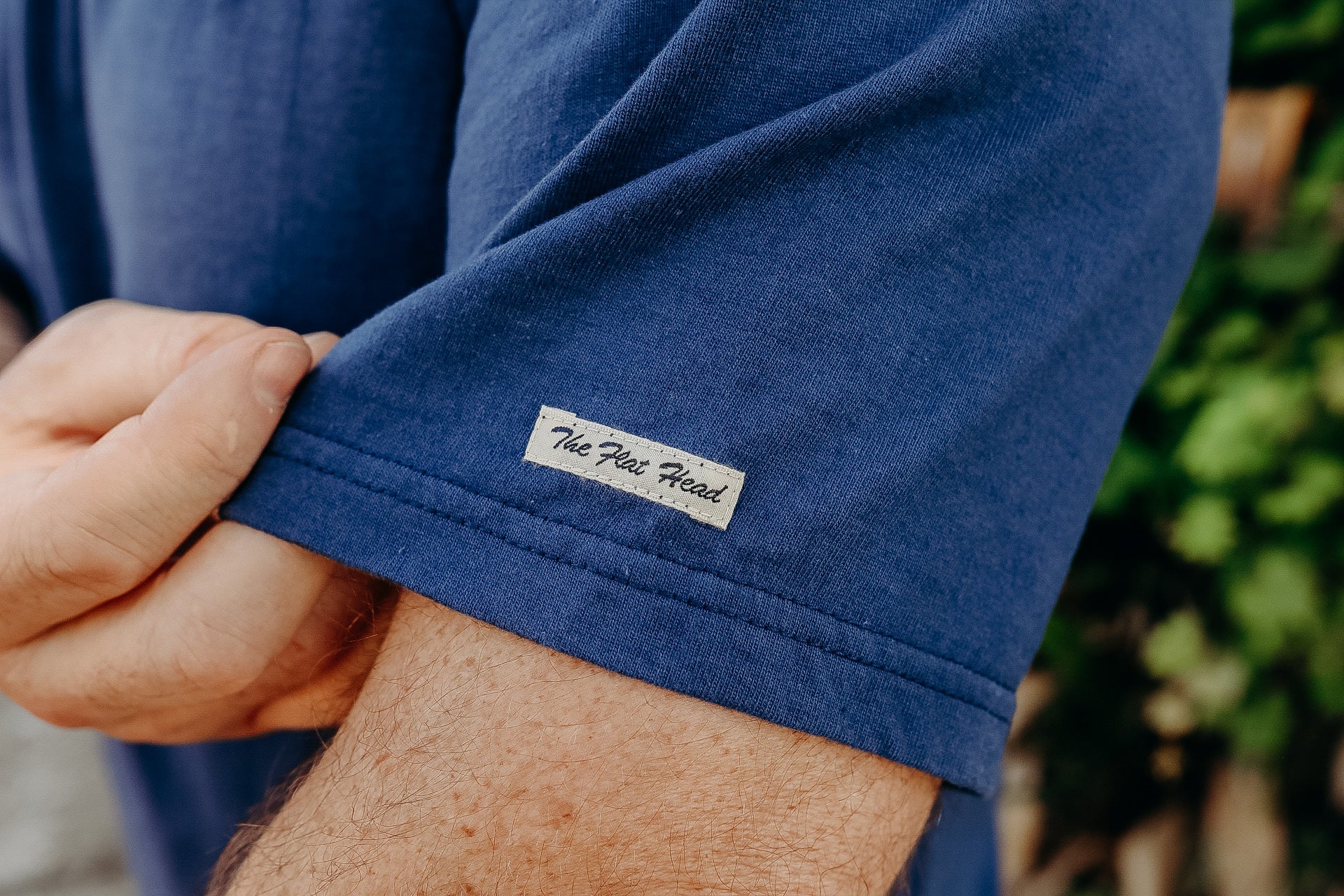 Close-up of a person's hand adjusting a blue short sleeve t-shirt with The Flat Head label on the hem