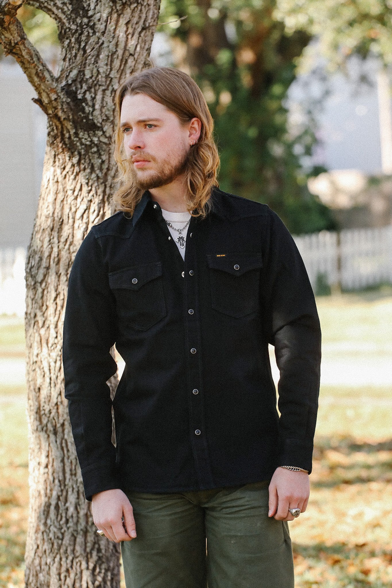 Man with long hair wearing a black button-up shirt and green pants standing outdoors by a tree