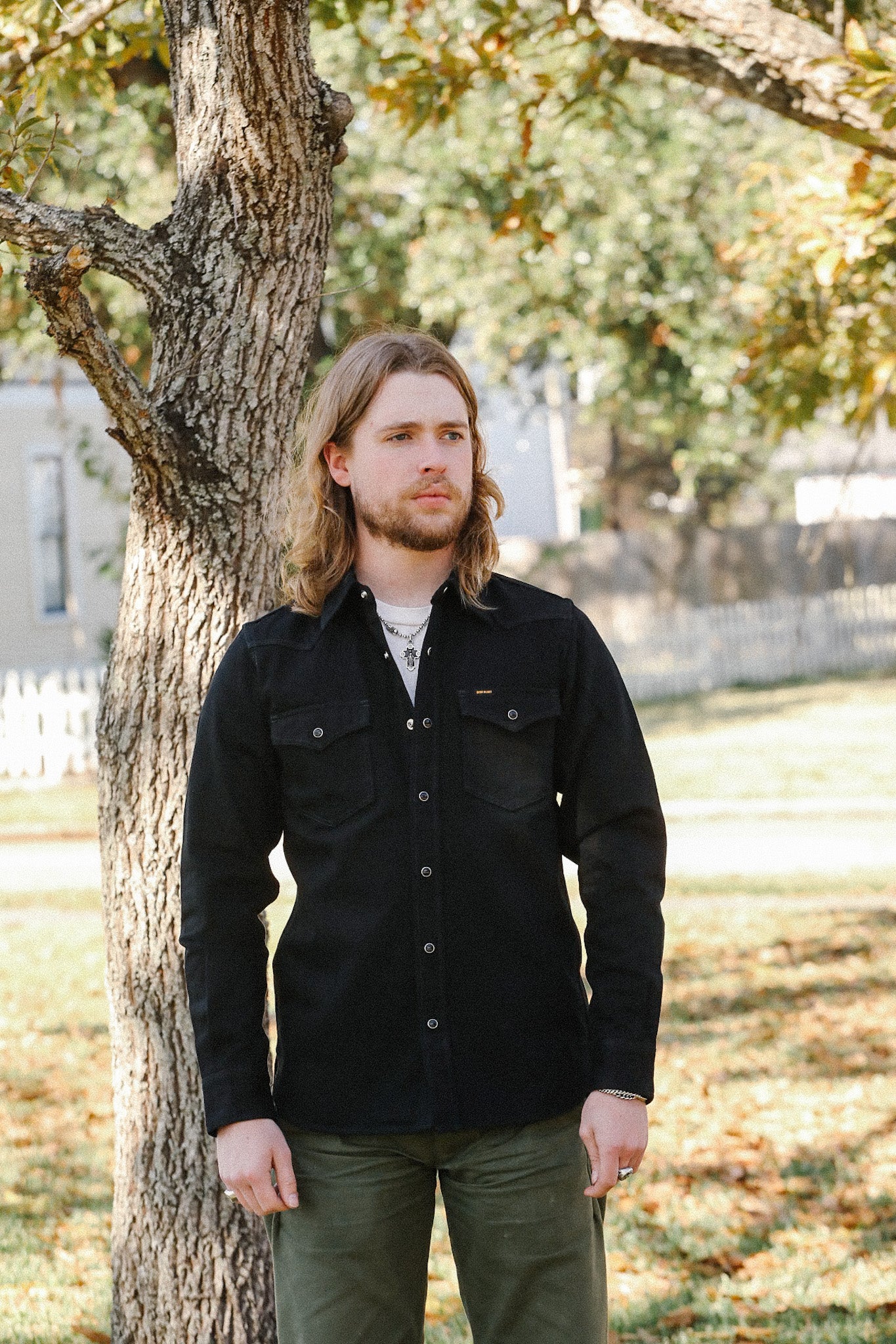 Man with long hair wearing black button-up shirt and green pants standing outdoors by tree