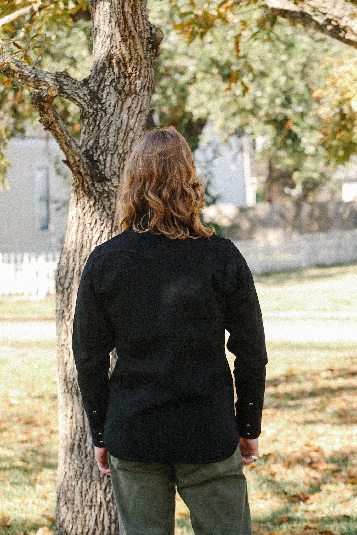 Person with long wavy hair wearing a black long sleeve shirt and olive pants standing outdoors by a tree