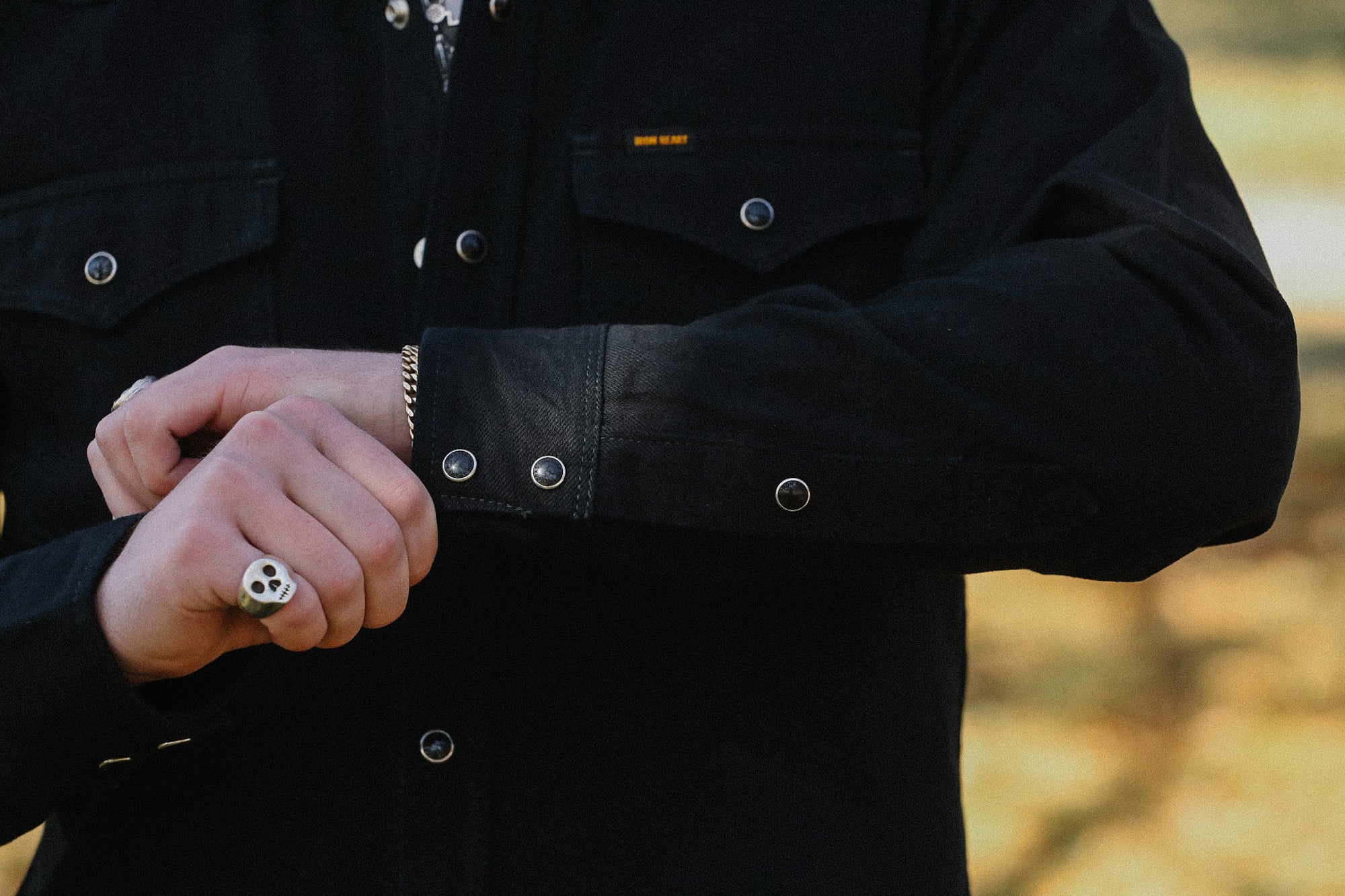 Close-up of person wearing black snap-button shirt with skull ring and silver bracelet adjusting cuff