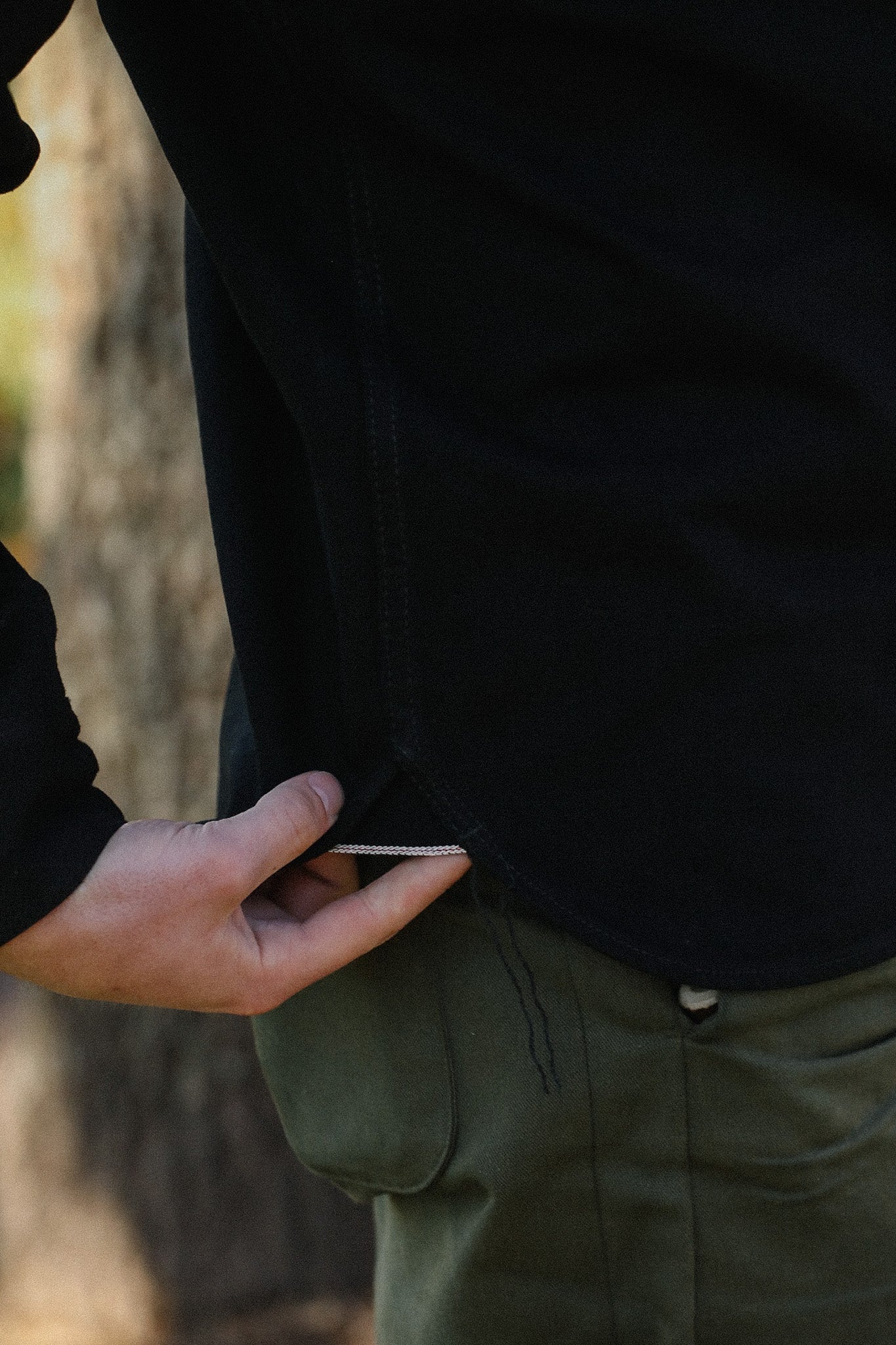 Close-up of hand lifting black shirt hem showing detailed stitching with green work pants pocket