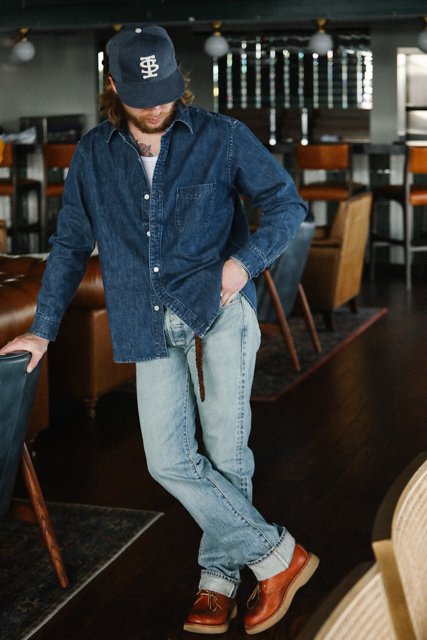 Man wearing denim shirt, light blue jeans, brown leather boots, and navy cap leaning on chair in stylish indoor setting