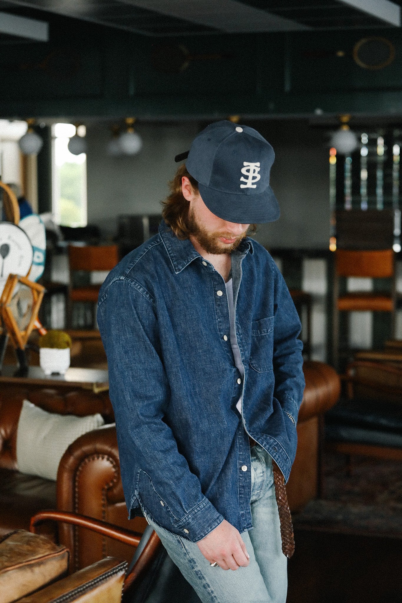 Man wearing a dark denim shirt and navy cap with white logo in a cozy vintage-style living room