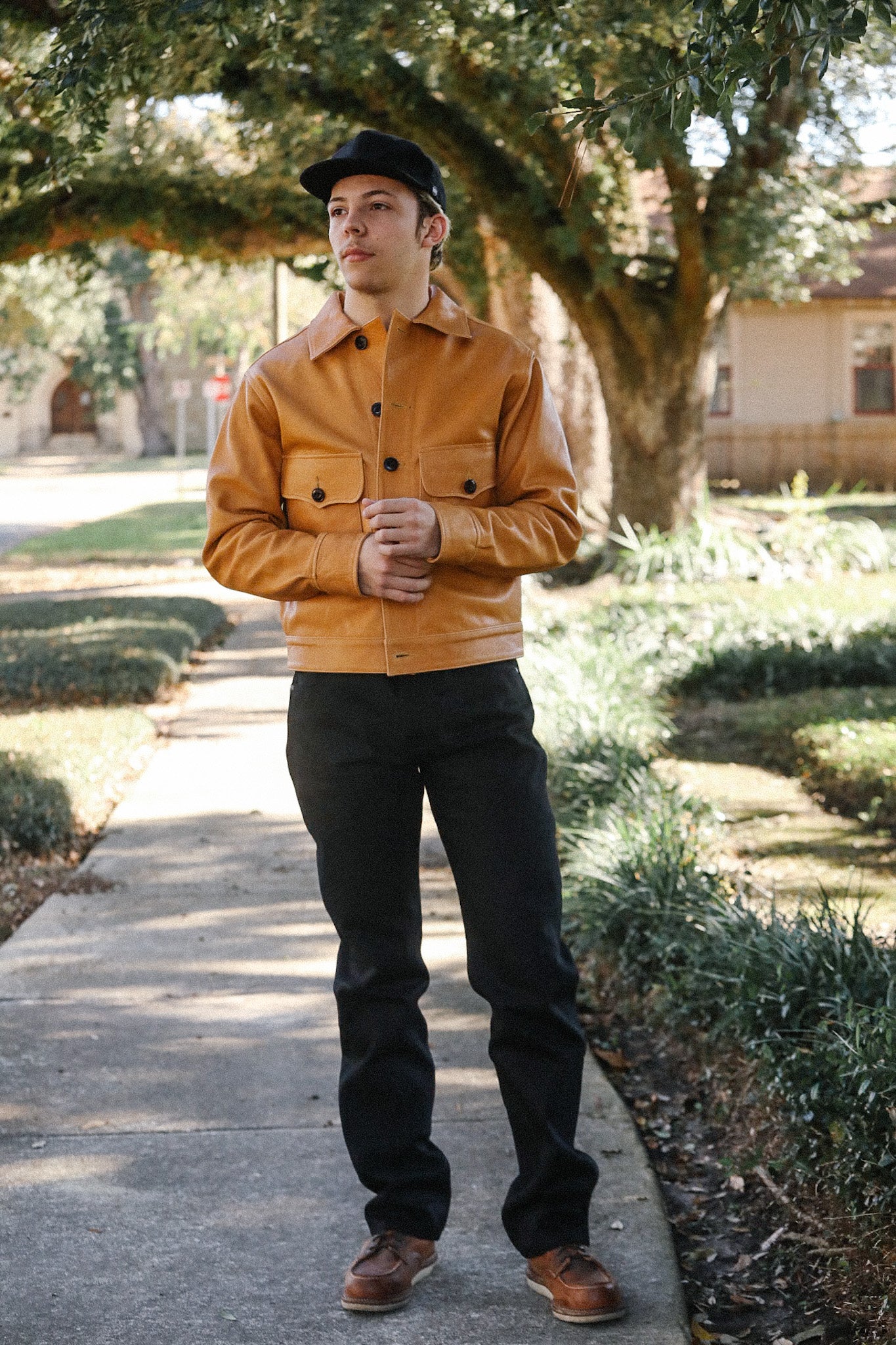 Man wearing tan leather jacket, black pants, brown shoes, and black cap standing on sunny sidewalk under trees