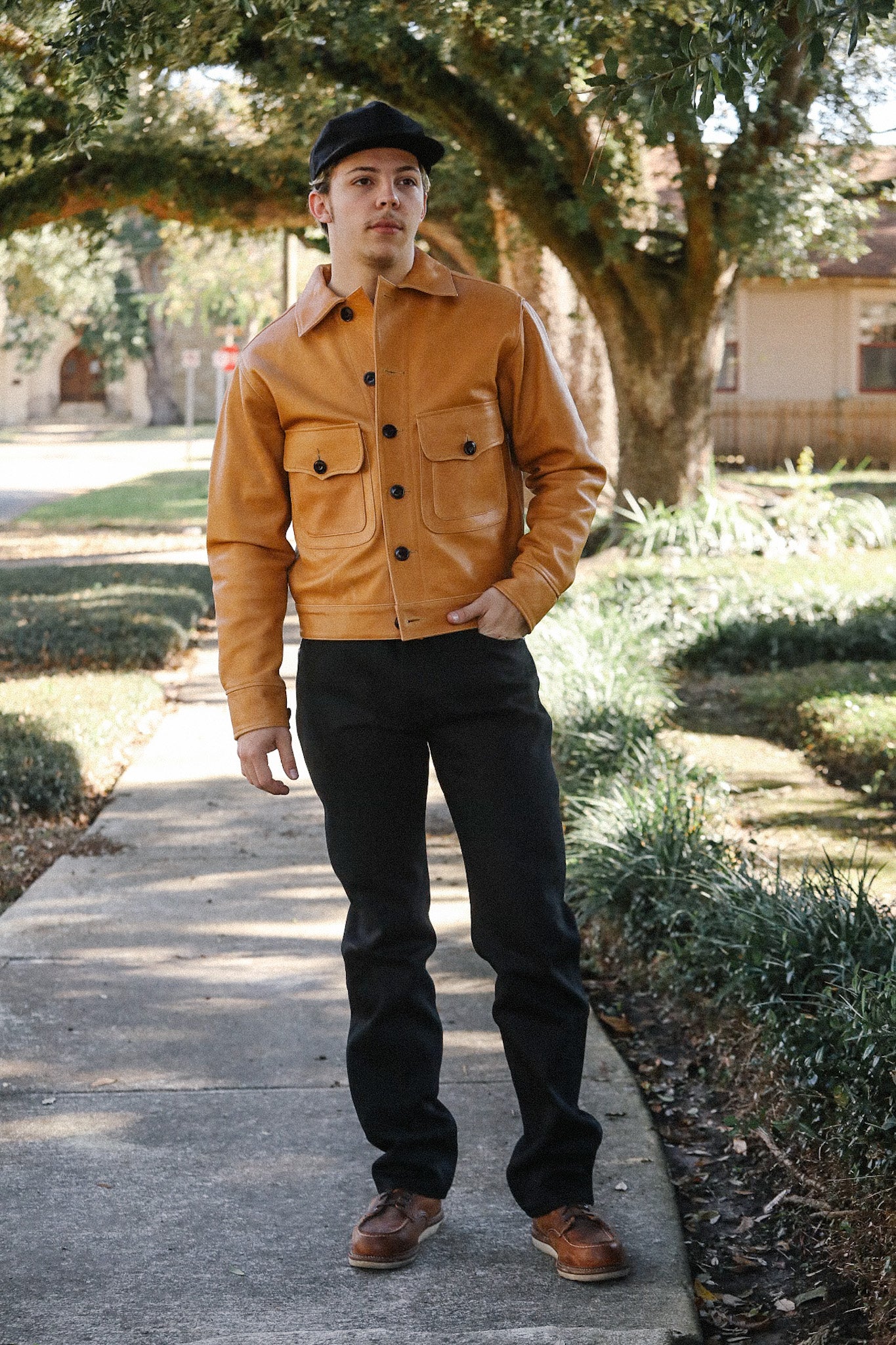 Man wearing tan leather jacket, black pants, brown shoes, and black cap standing on sidewalk outdoors