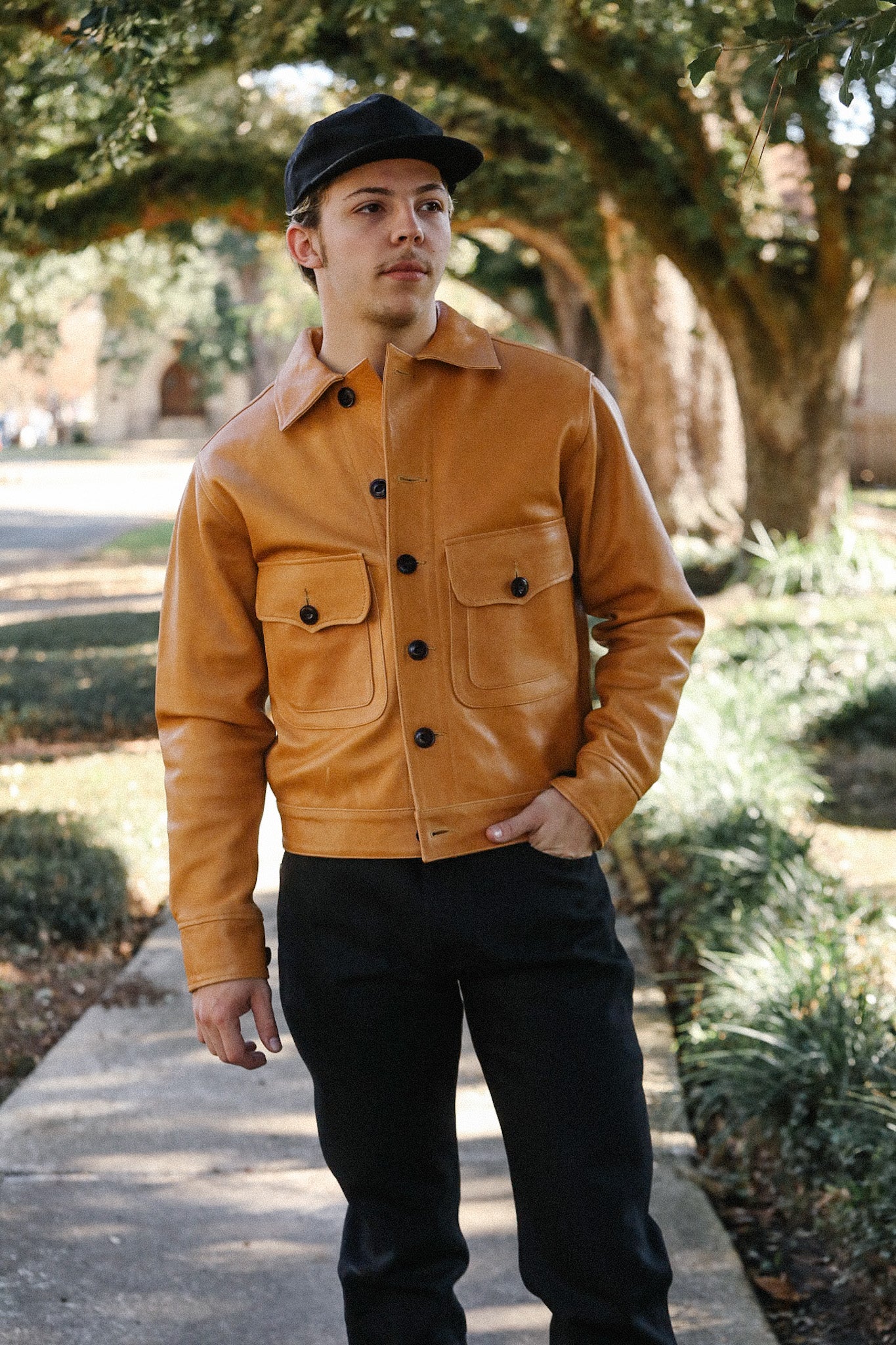 Man wearing a tan leather jacket with black buttons and pockets, black pants, and a black cap outdoors