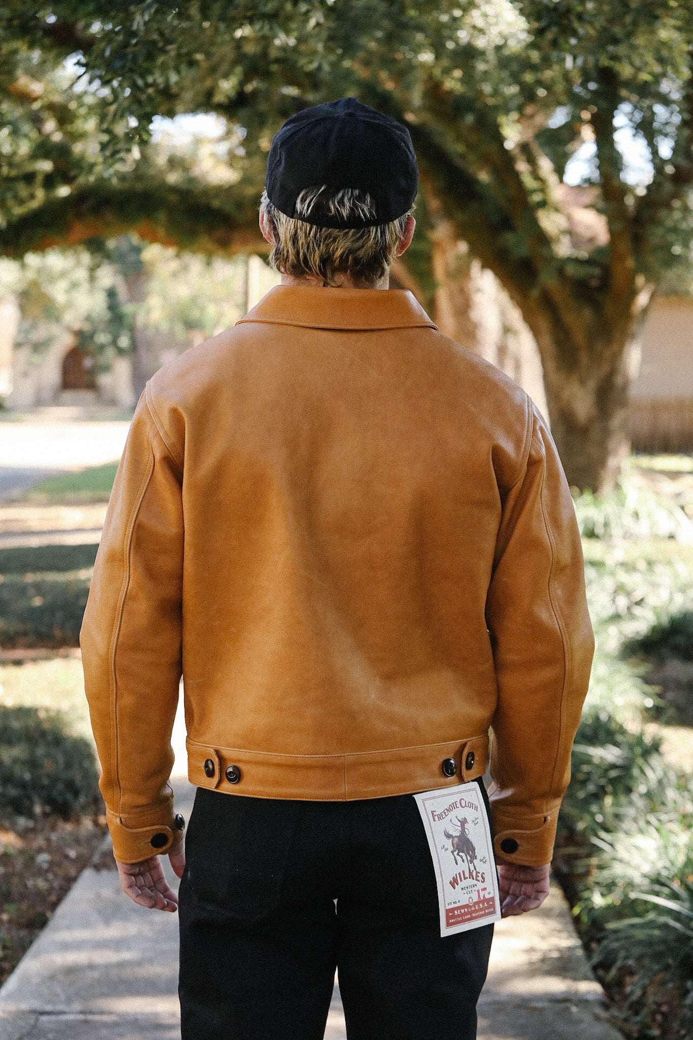 Man wearing a brown leather jacket and black cap walking outdoors with trees and path in background