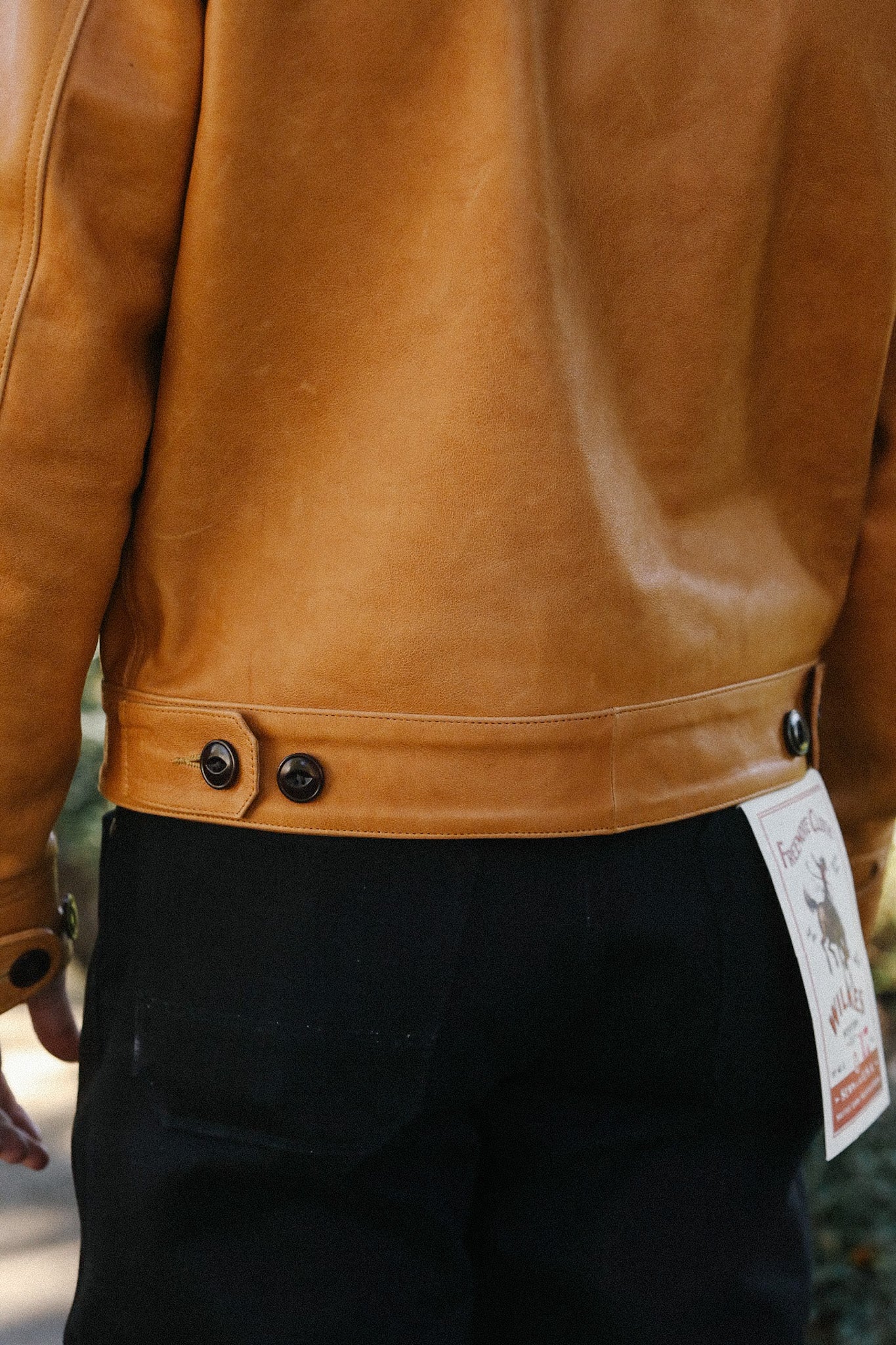 Back view of person wearing tan leather jacket and black pants with tag, outdoor setting
