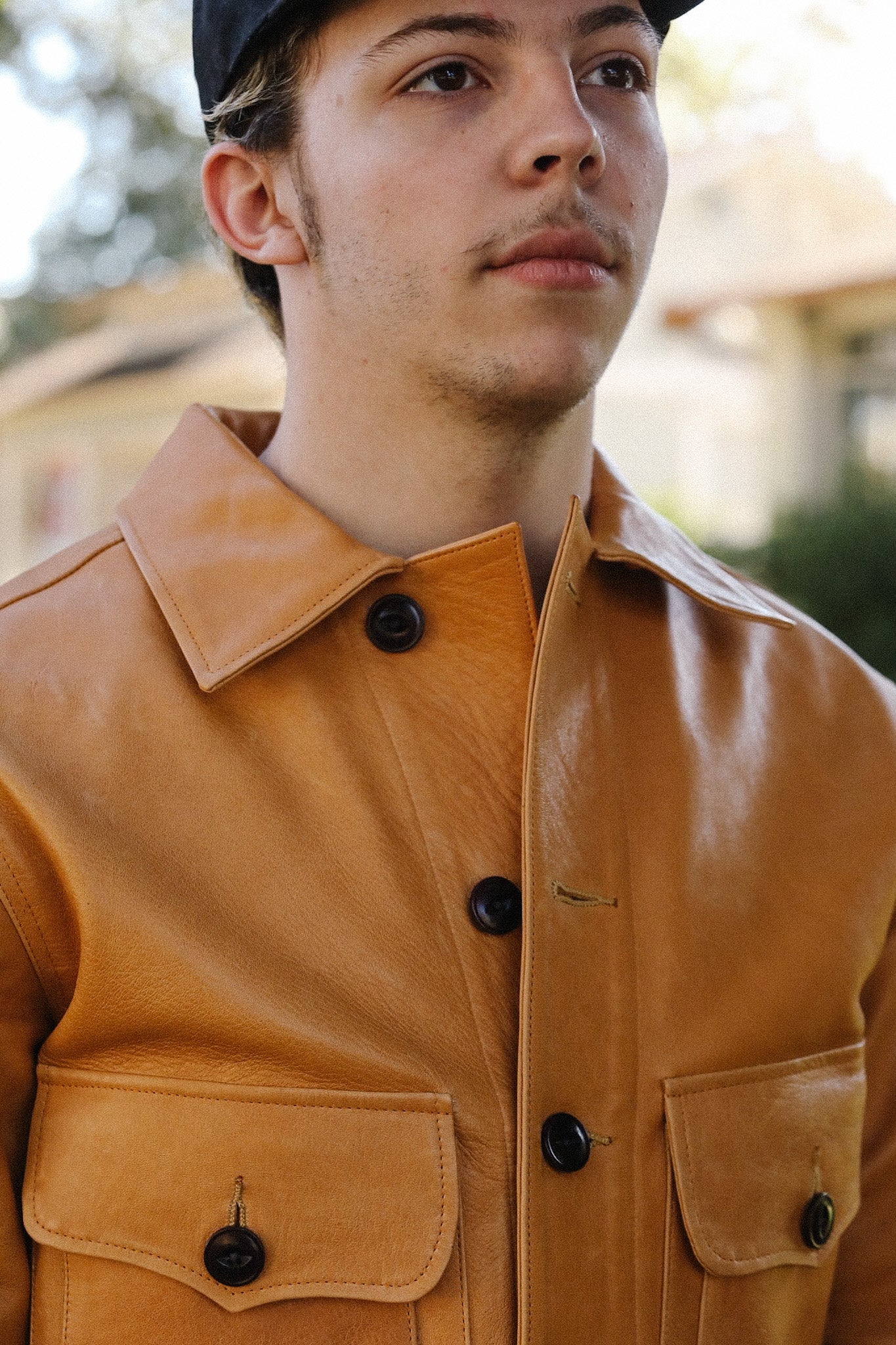 Close-up of a young man wearing a tan leather jacket with large black buttons and flap pockets outdoors