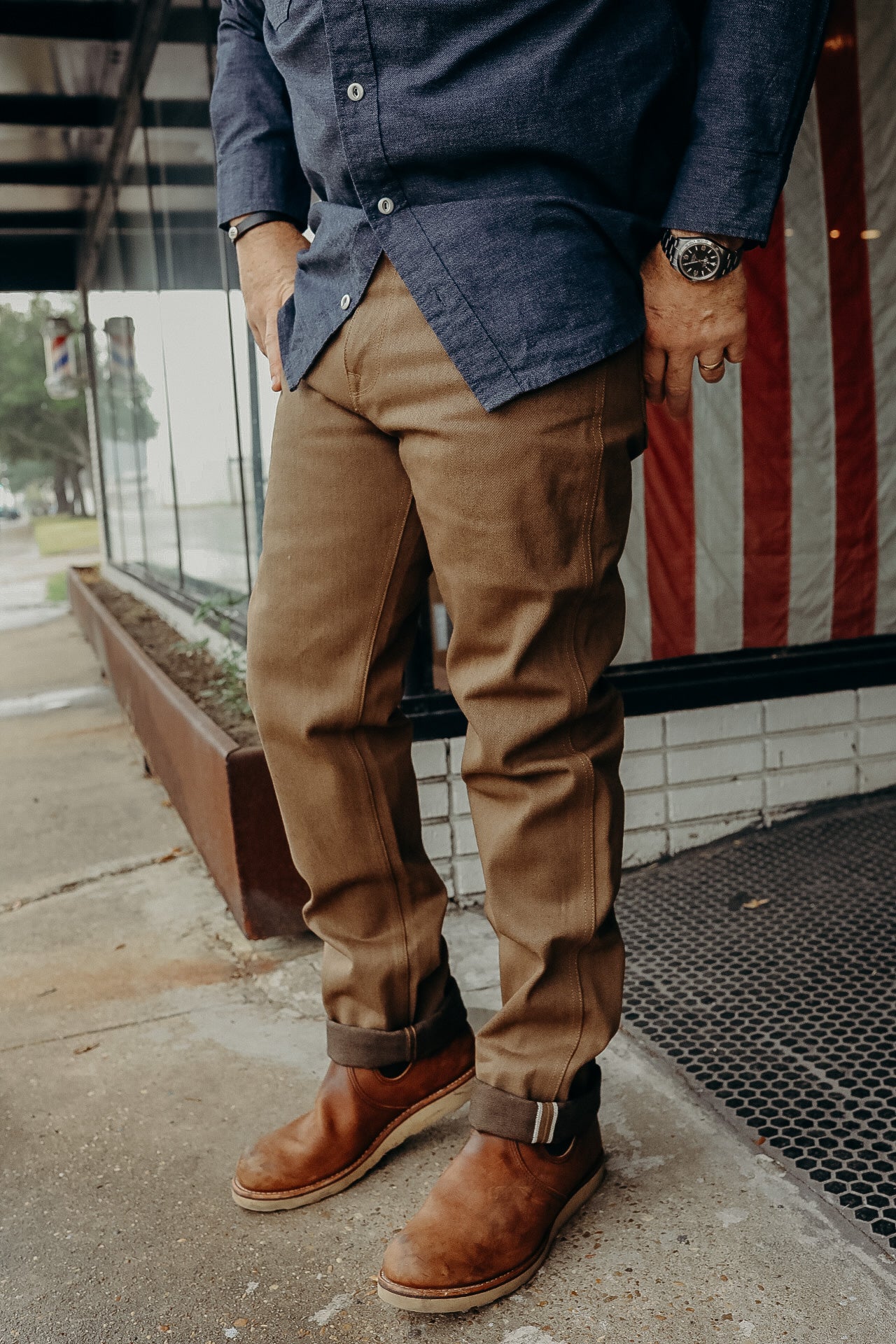 Man wearing brown work pants and brown leather boots standing outside a storefront with an American flag backdrop