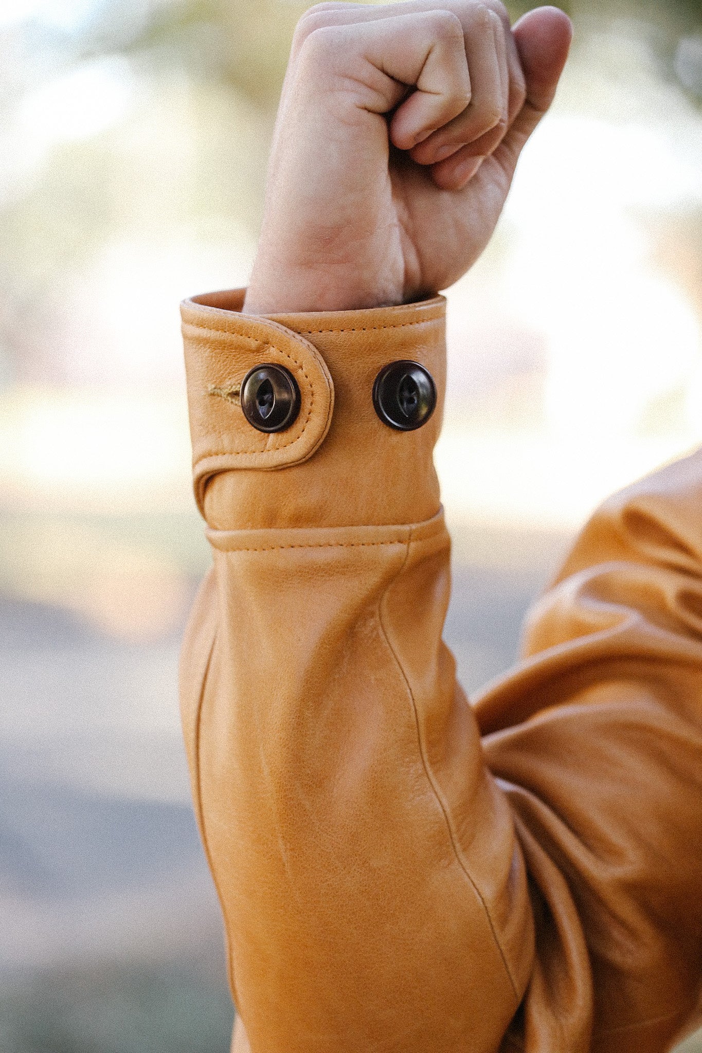 Close-up of a fist raised inside a tan leather jacket cuff with two dark buttons, outdoors