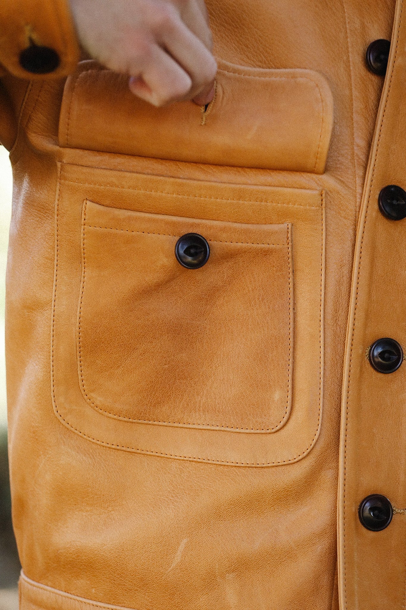 Close-up of a person’s hand opening the top flap of a tan leather jacket pocket with black button details