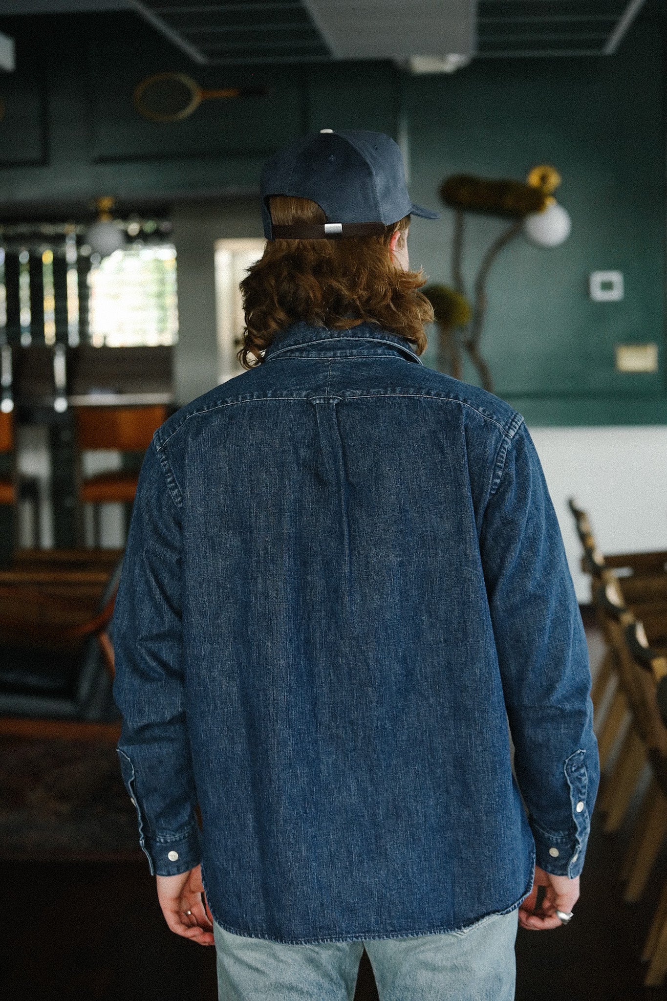 Man wearing denim shirt and navy cap standing indoors in a rustic room with wooden chairs
