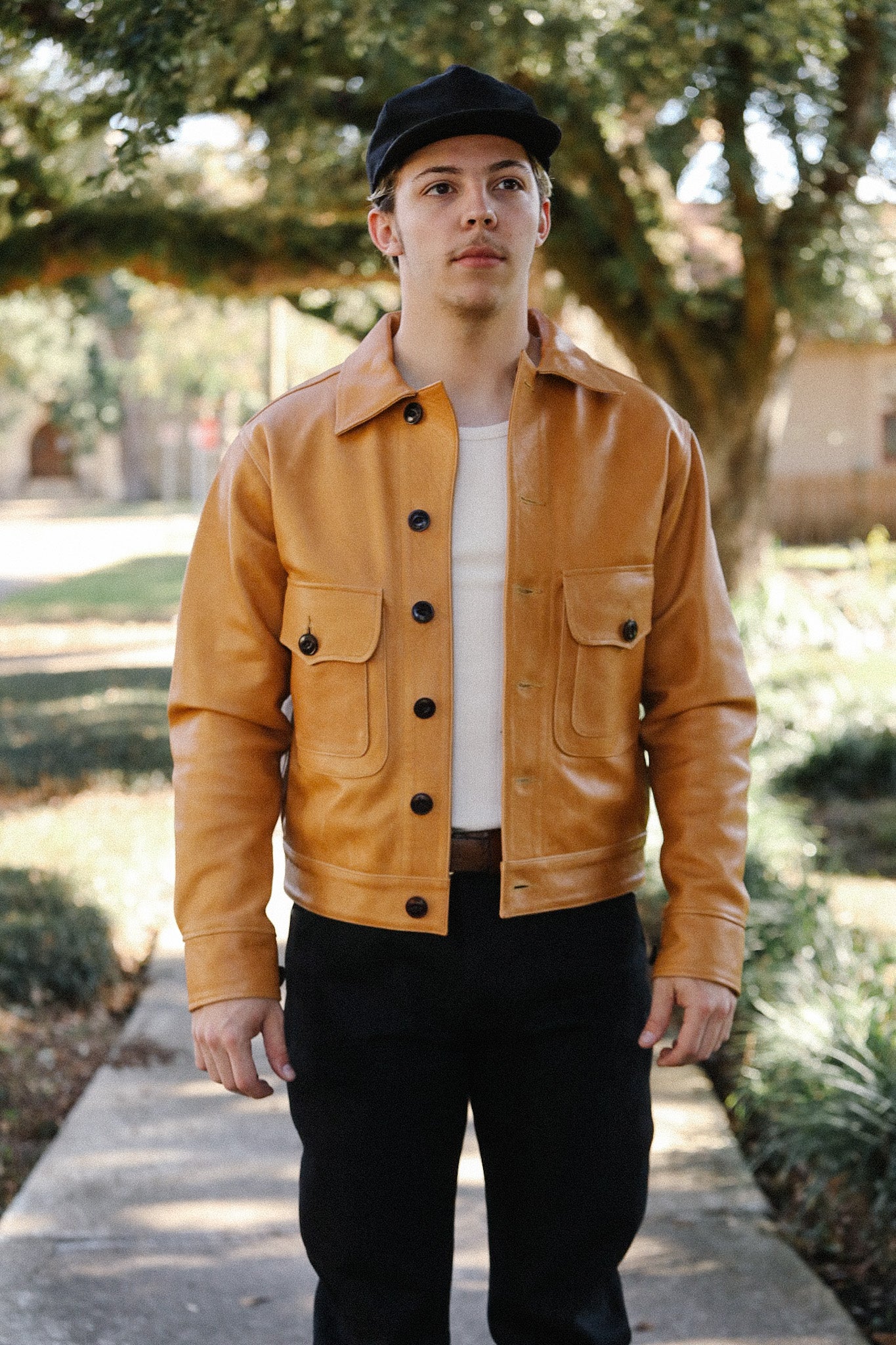 Man wearing tan leather jacket, black pants, white shirt, and black cap standing outdoors on pathway