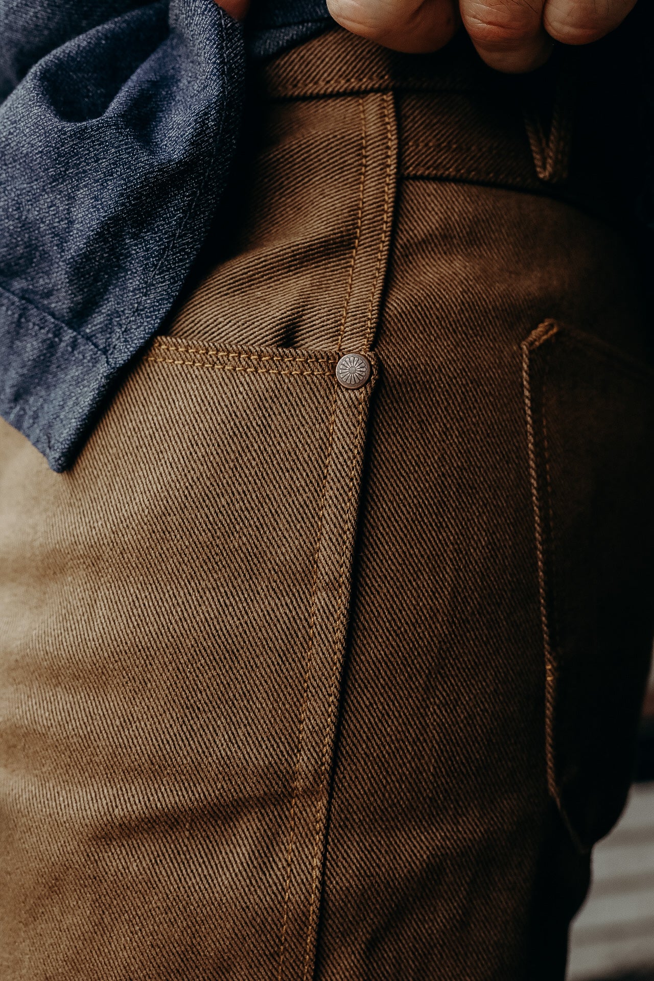 Close-up of brown rugged denim pants pocket with detailed stitching and metal rivet, worn with blue denim shirt