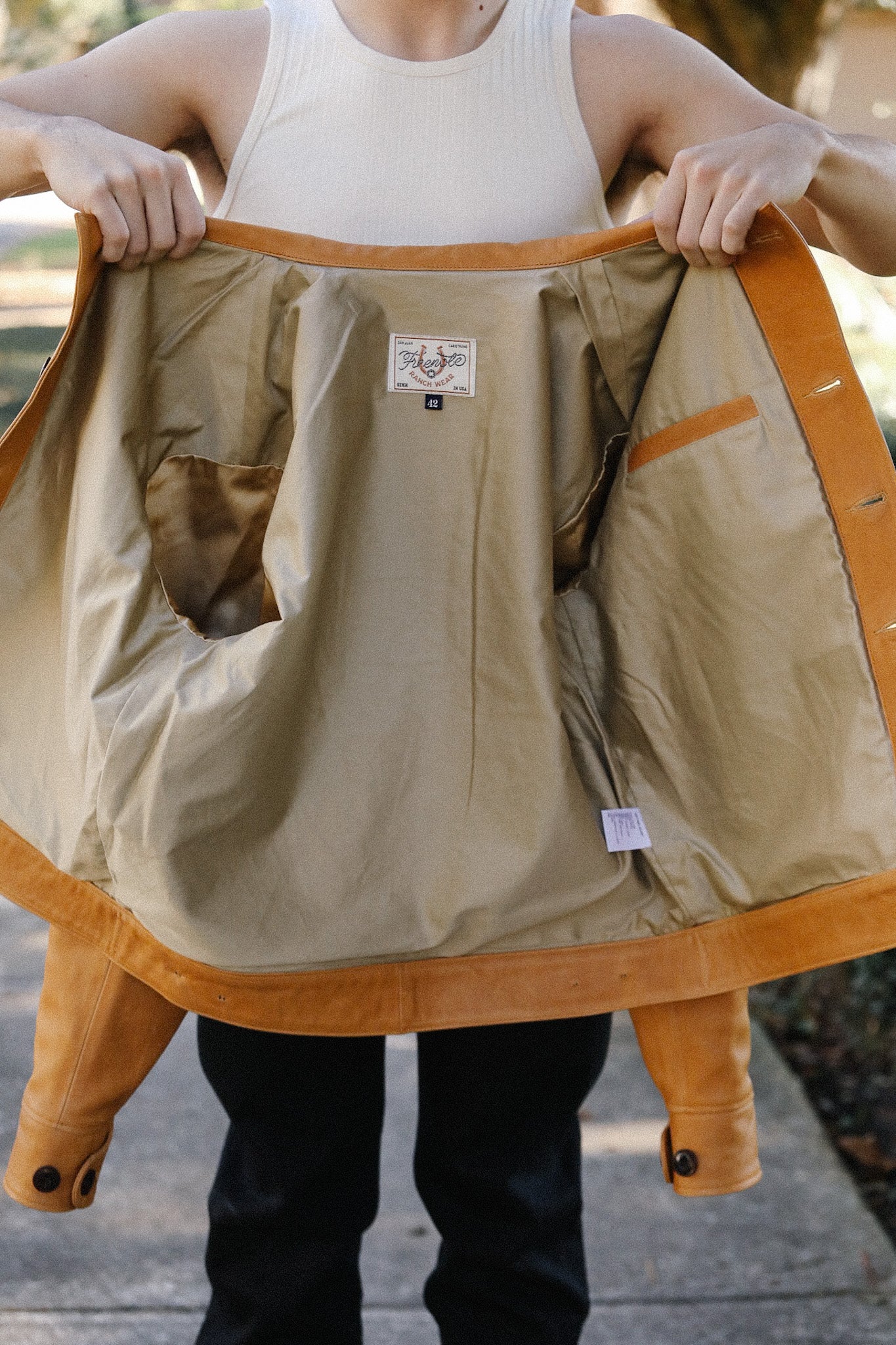 Person holding open a tan leather jacket showing beige inner lining with pockets and label inside