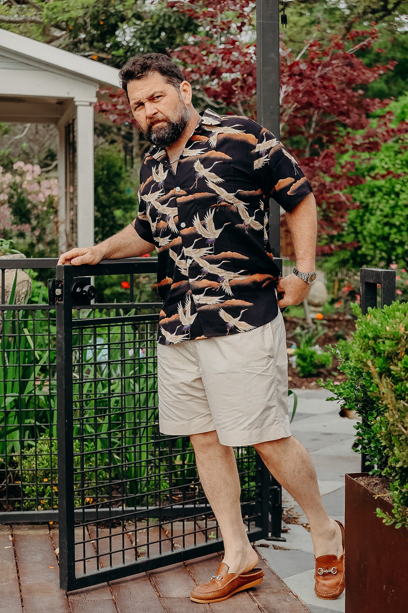 man in black bird print shirt and beige shorts opening garden gate in lush greenery