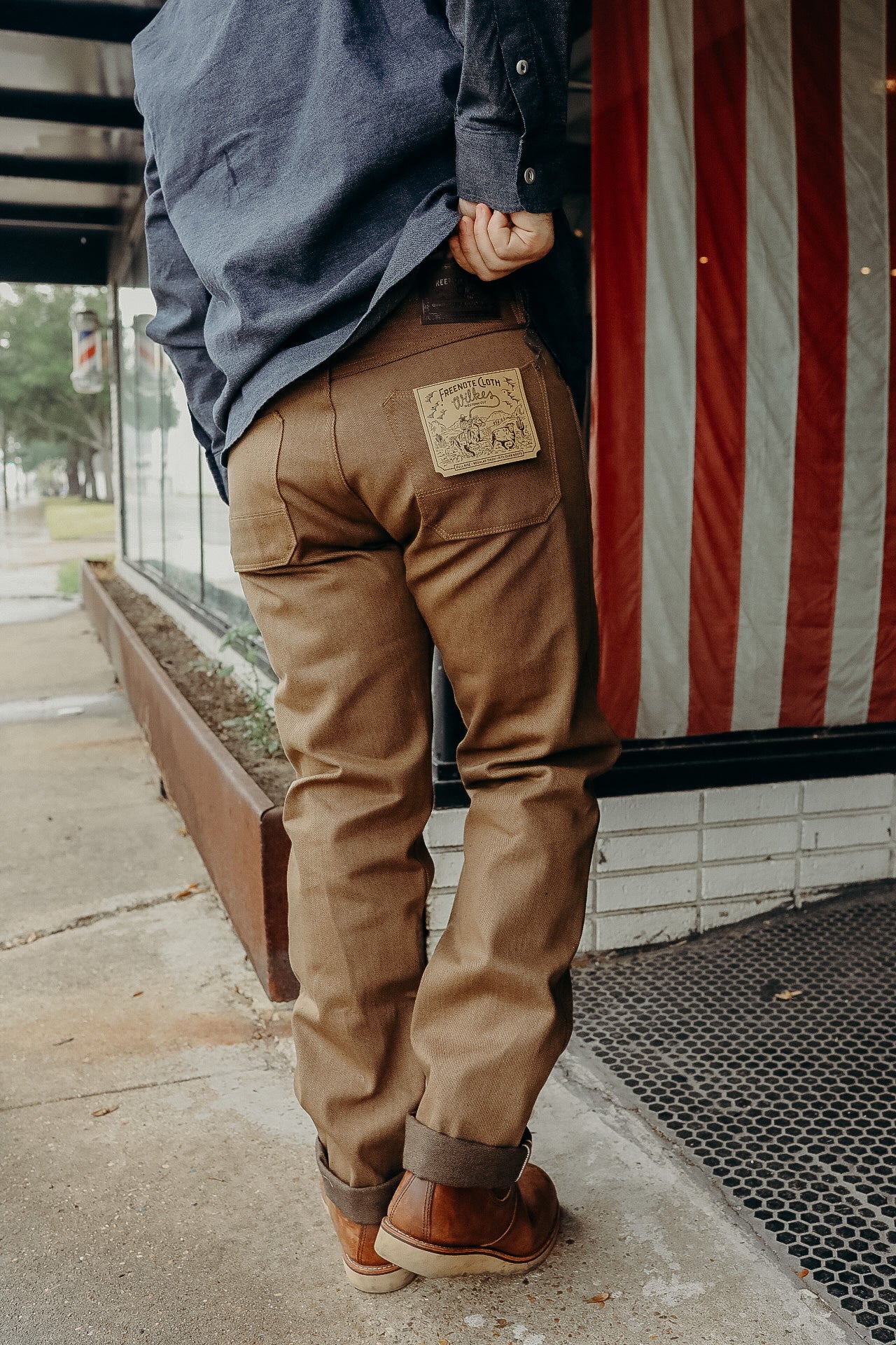 Man wearing brown work pants with label and blue shirt, standing near American flag outside building