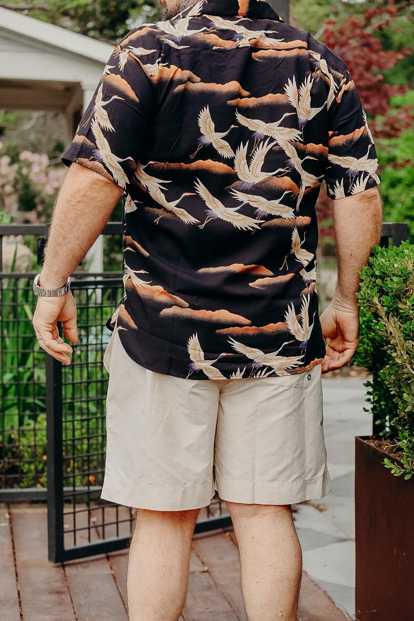 Man wearing black Hawaiian shirt with crane bird print and beige shorts outdoors near garden gate