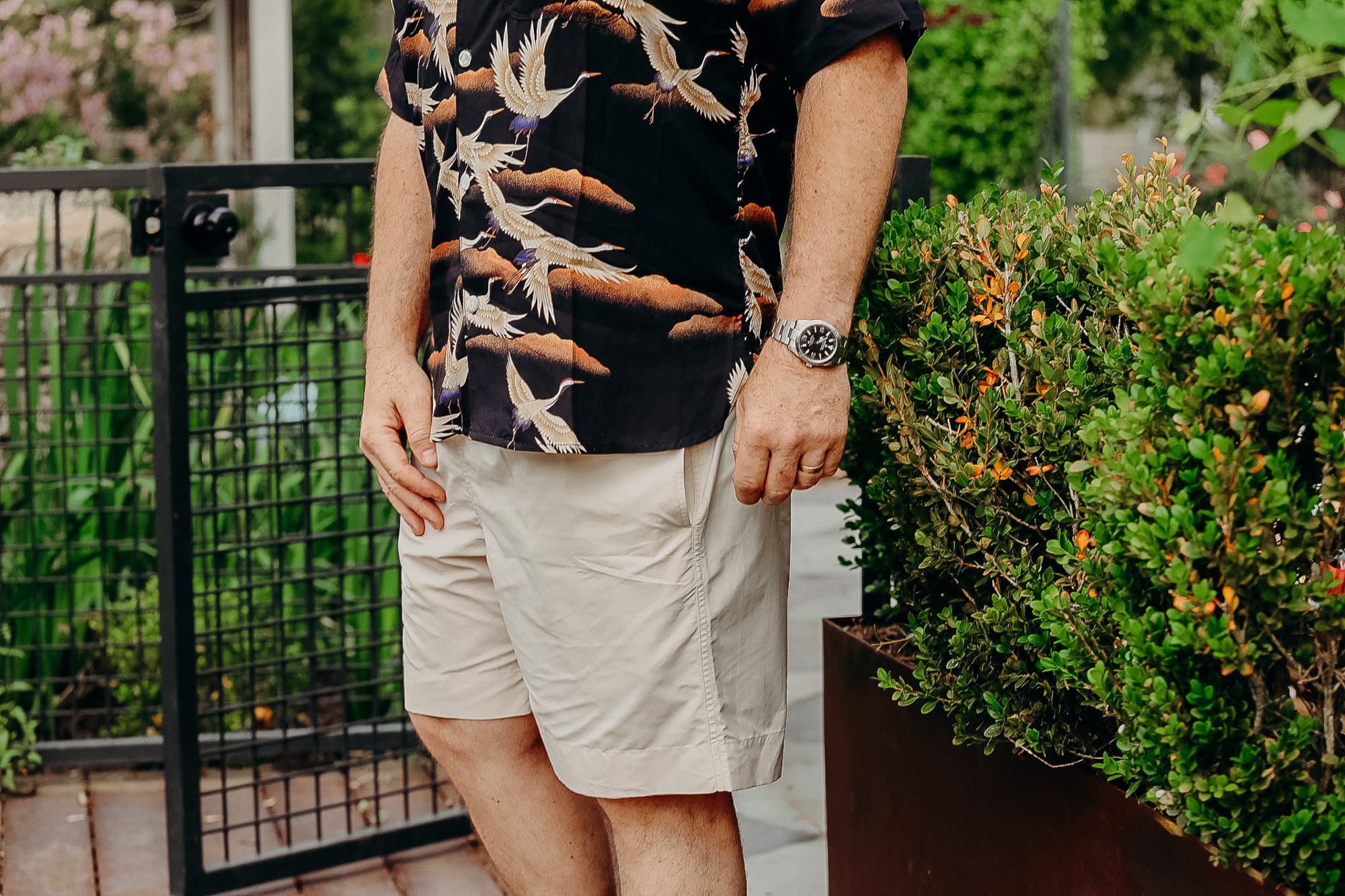 Man wearing tropical black shirt with crane print and beige shorts standing by green bushes