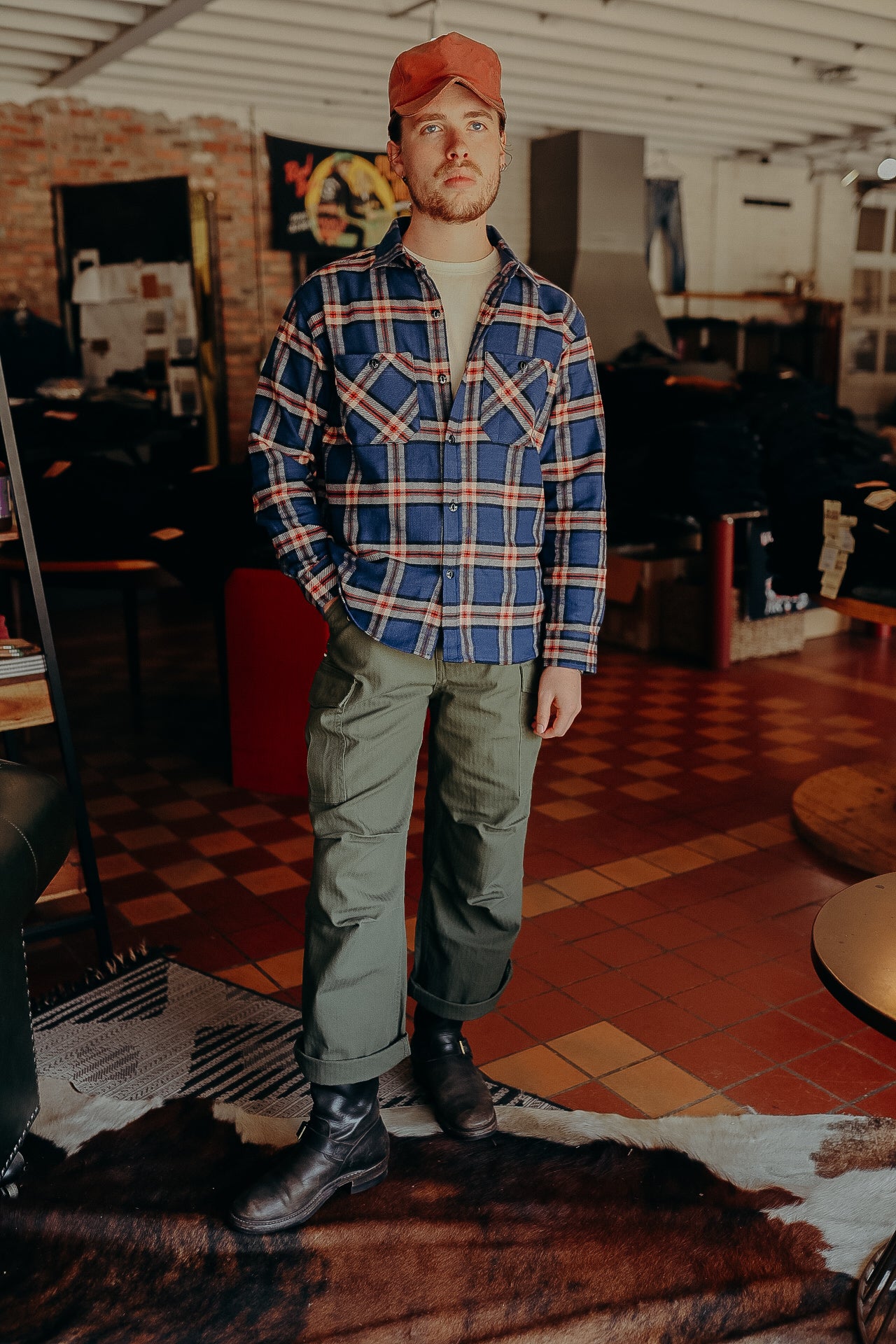 Man in red cap, blue plaid shirt, olive cargo pants, and black boots standing on cowhide rug in rustic store