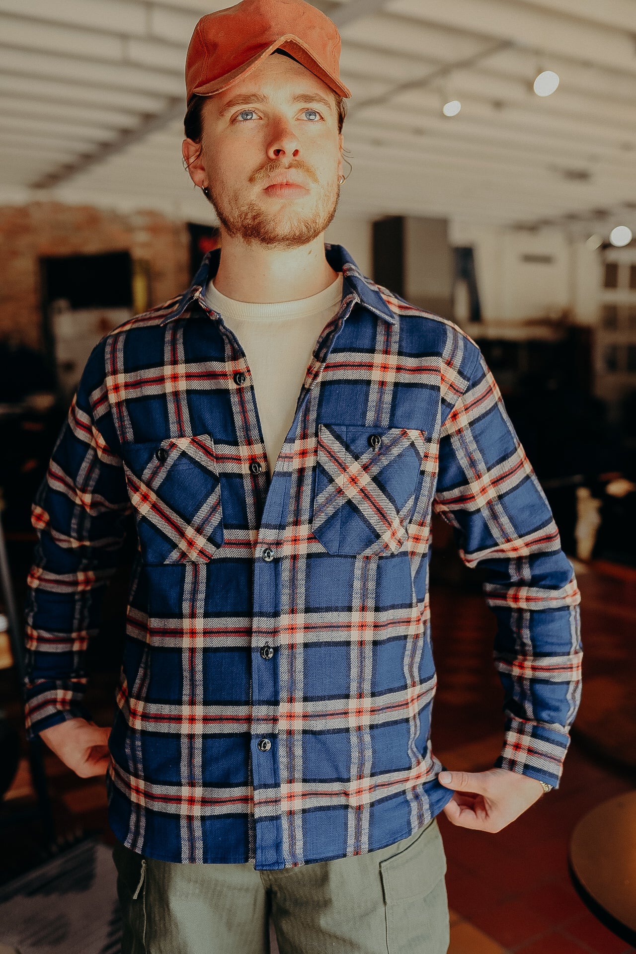 Man wearing blue plaid flannel shirt, beige t-shirt, red cap, and green pants indoors at Iron Shop Provisions