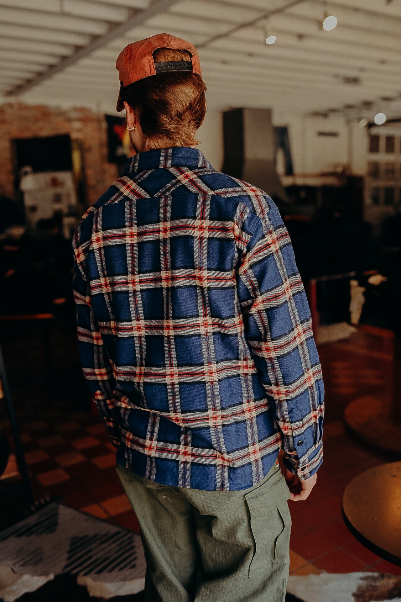 Back view of person wearing blue and red plaid shirt, olive green cargo pants, and orange cap indoors