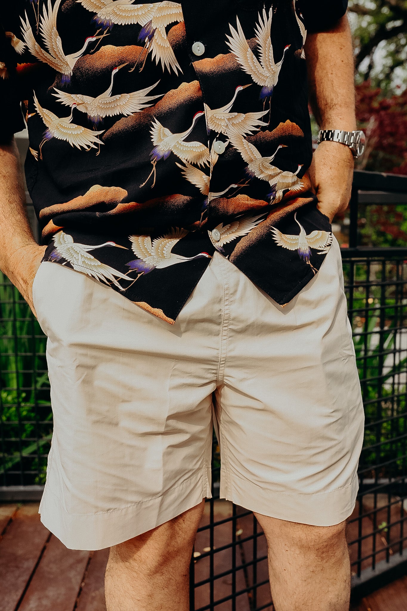 Man wearing beige shorts and black button-up shirt with crane bird print, standing outdoors with hands in pockets