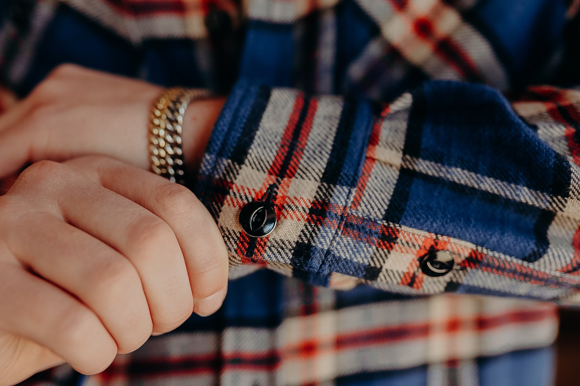 Close-up of hands adjusting sleeve cuff on blue and red plaid shirt with gold chain bracelet