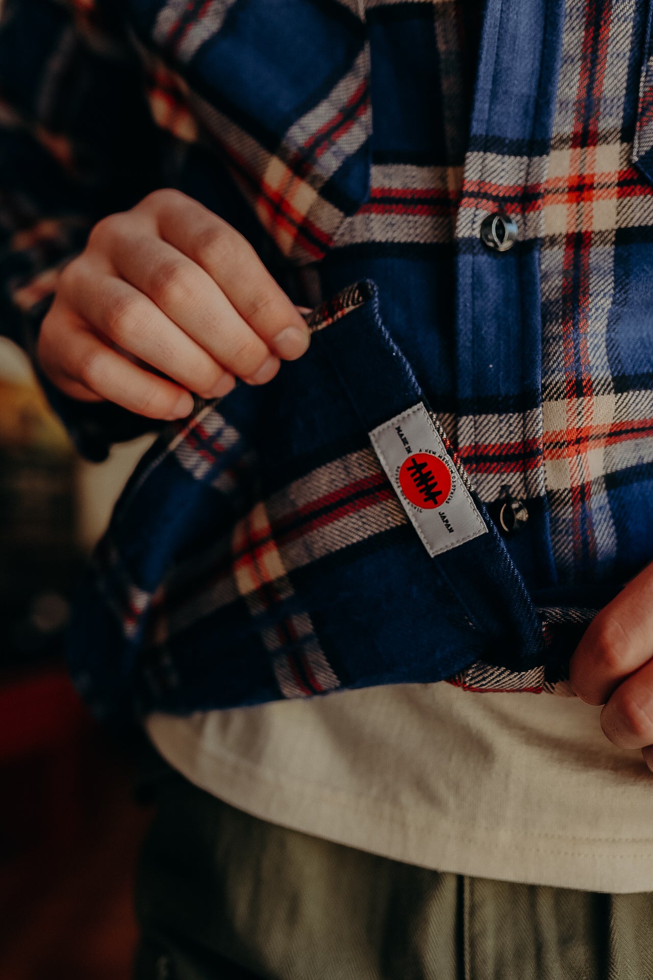 Close-up of a person wearing a blue and red plaid shirt with a visible tag saying Made in Japan, holding the shirt hem
