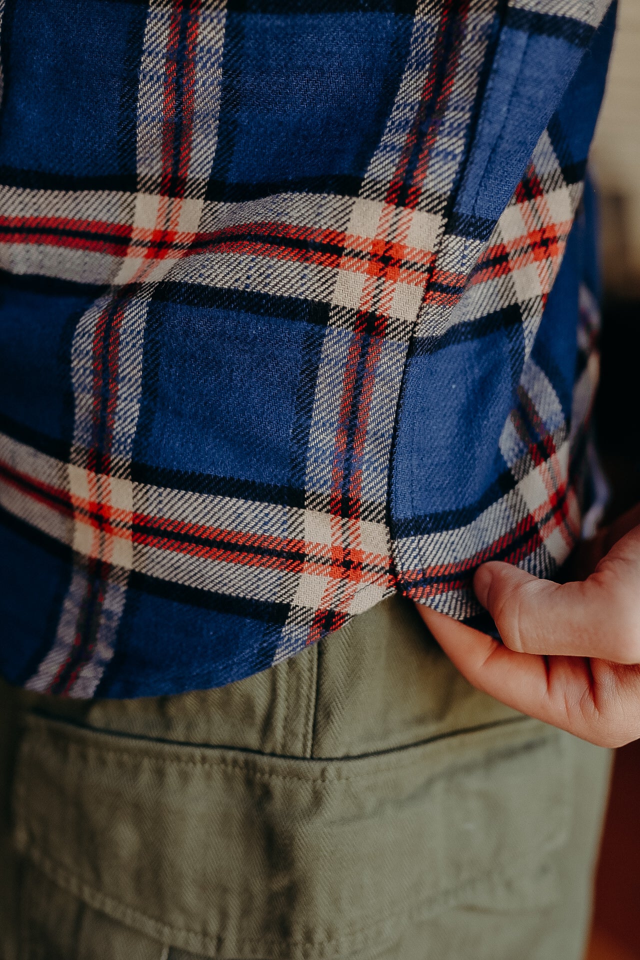 Close-up of a hand holding the hem of a blue plaid flannel shirt with red and white stripes, paired with green pants