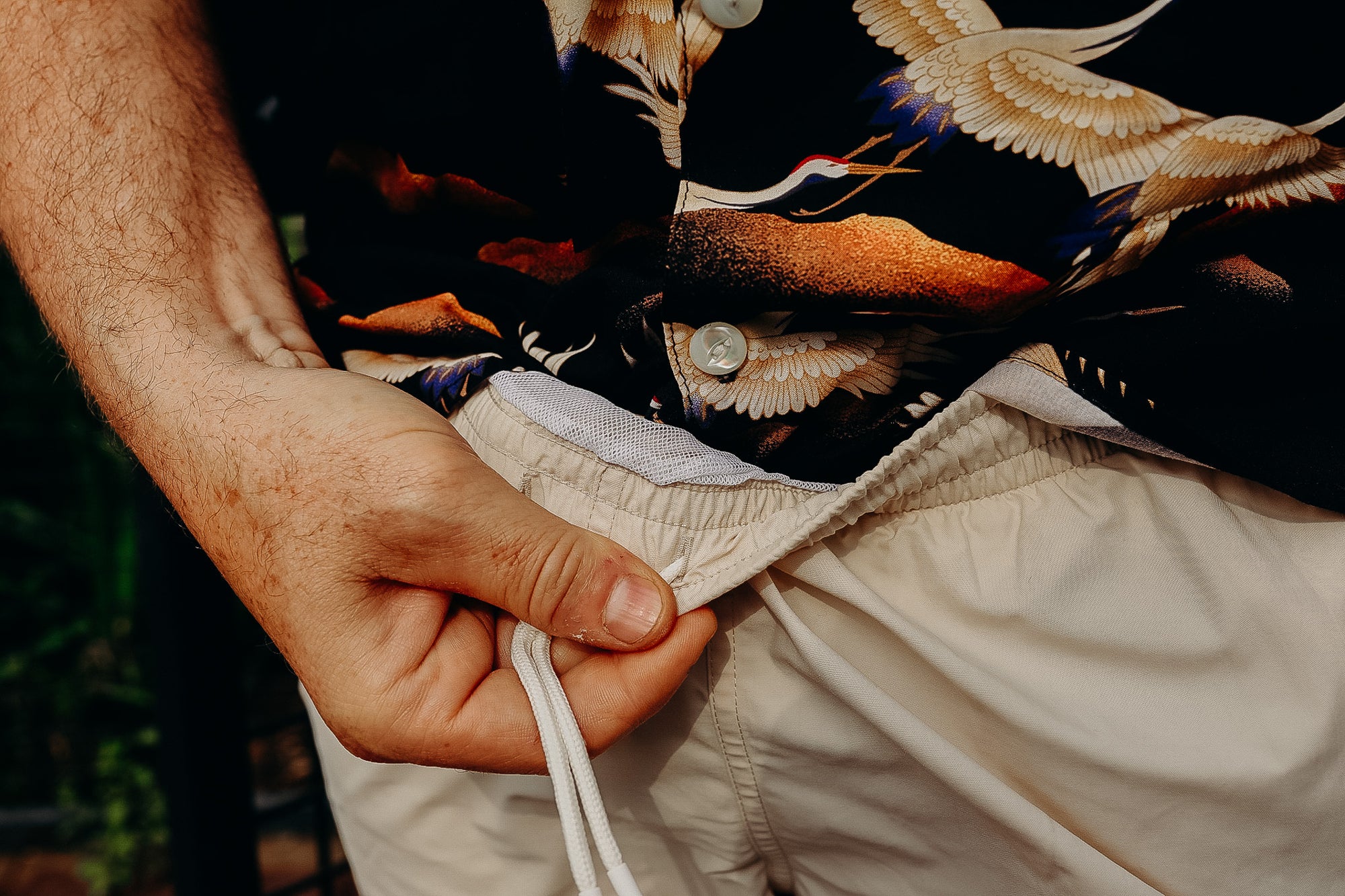 Close-up of hand adjusting beige drawstring shorts paired with a black tropical crane print shirt