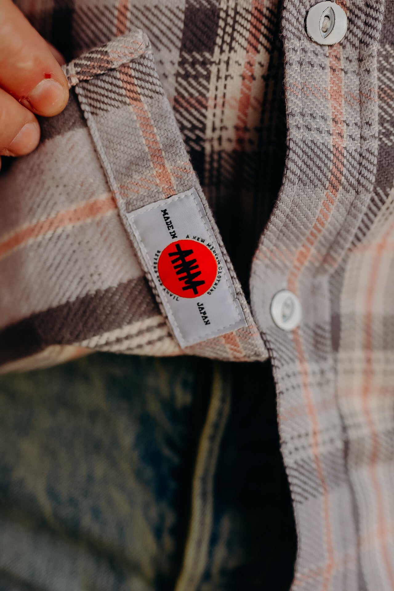 Close-up of hand holding a beige and orange plaid shirt with a Made in Japan label from Iron Shop Provisions
