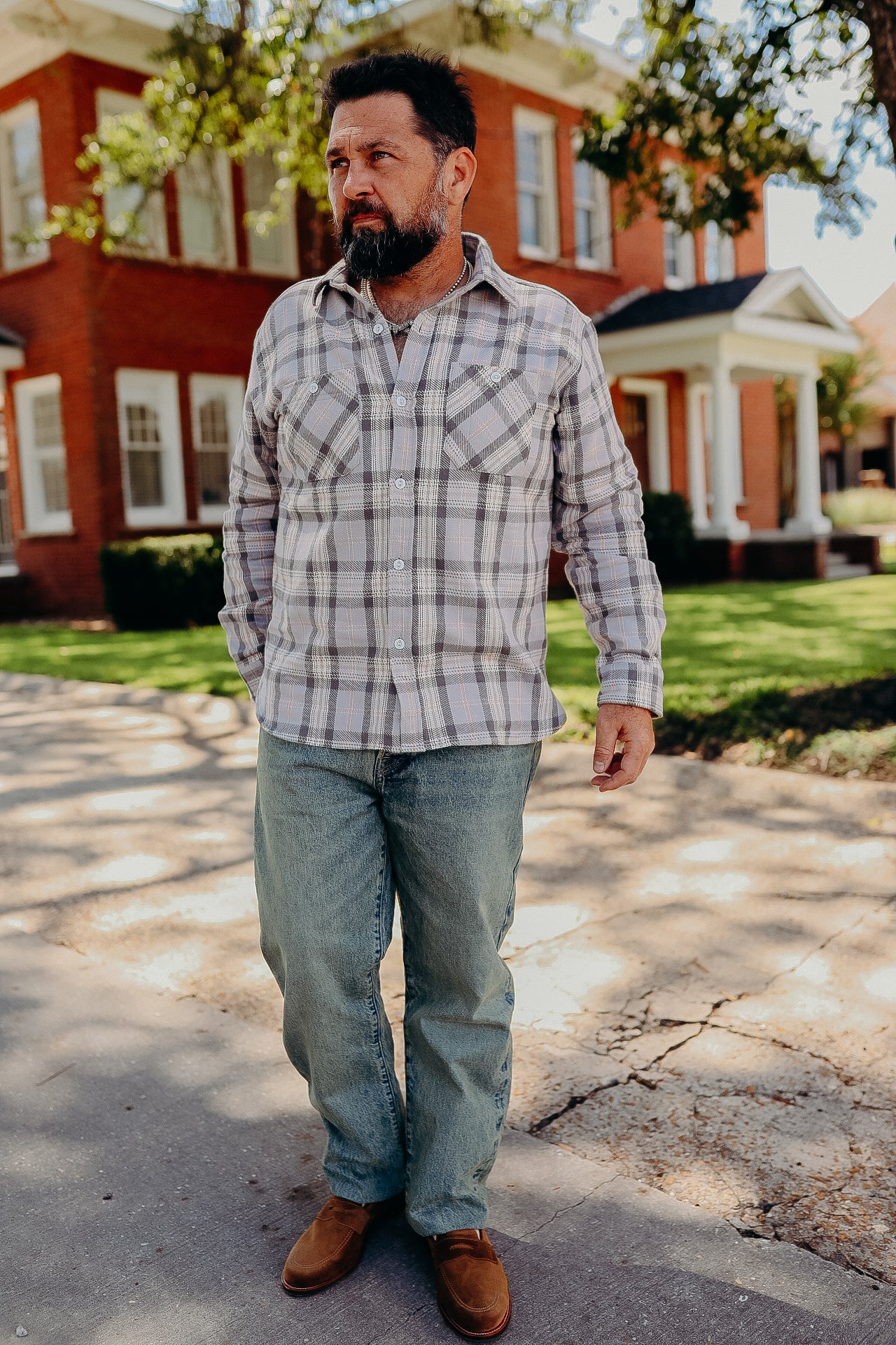 Bearded man wearing plaid shirt, worn jeans, and brown shoes walking on sunny sidewalk with red brick house background