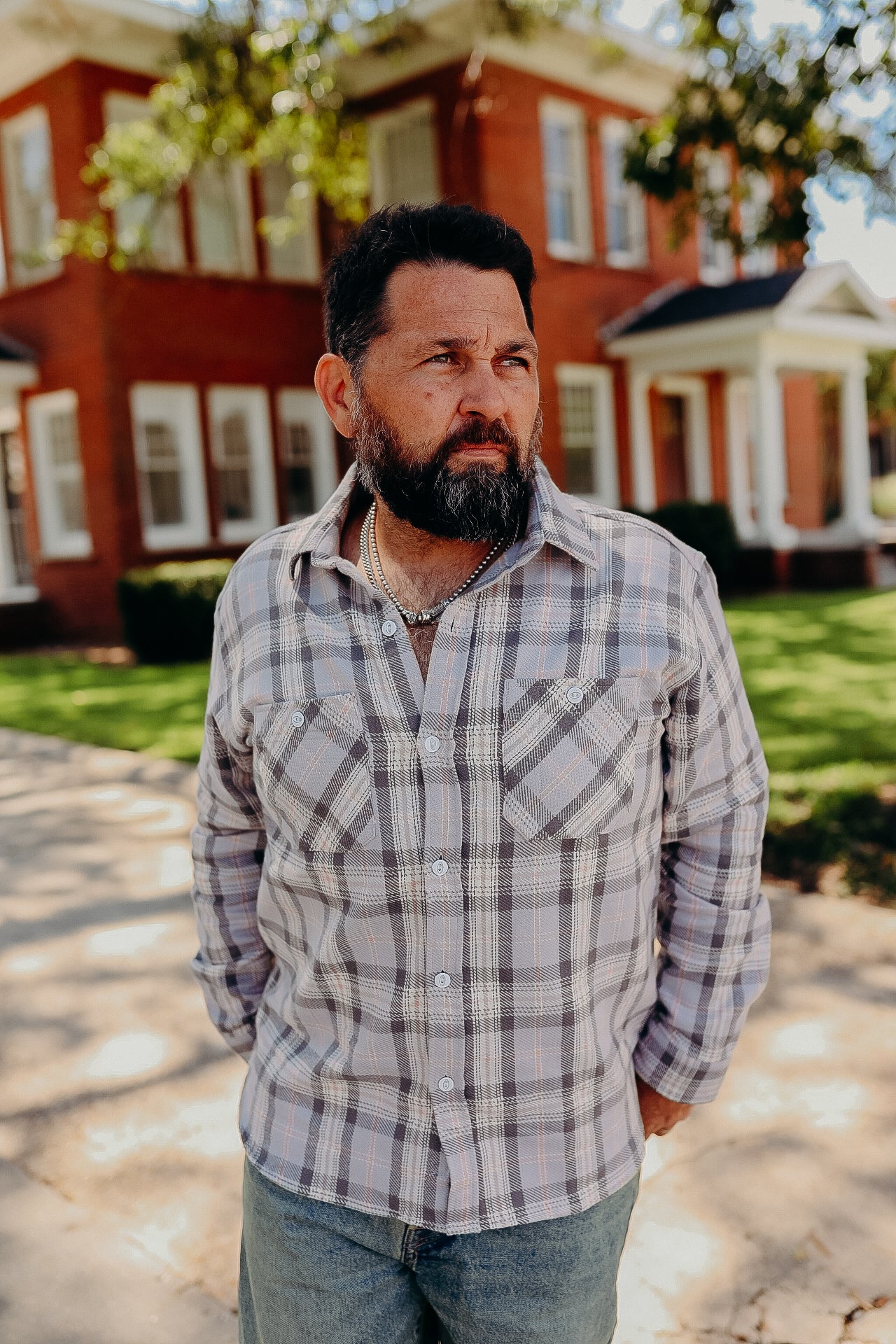 Bearded man in plaid button-up shirt and jeans standing outdoors with a brick house background